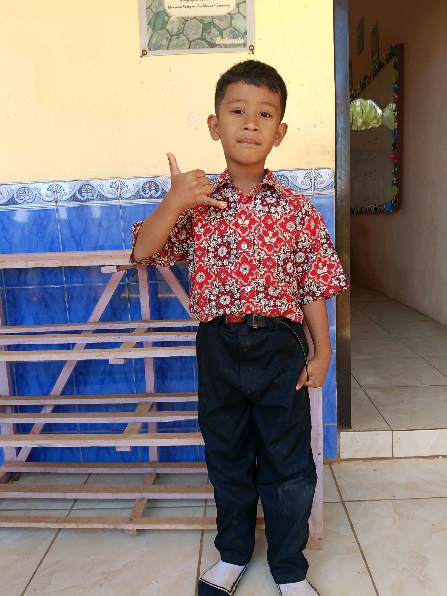 A young boy stands outside on a sunny day. He is wearing a red and white patterned shirt and black pants. His right hand is raised in a "shaka" gesture, and his left hand is in his pocket. Behind him, there is a blue tiled wall and a wooden shelf. To his right, a doorway leads into a darker interior. The ground is tiled with light-colored ceramic tiles. The image appears to have been taken in Gunungwungkal, Indonesia.