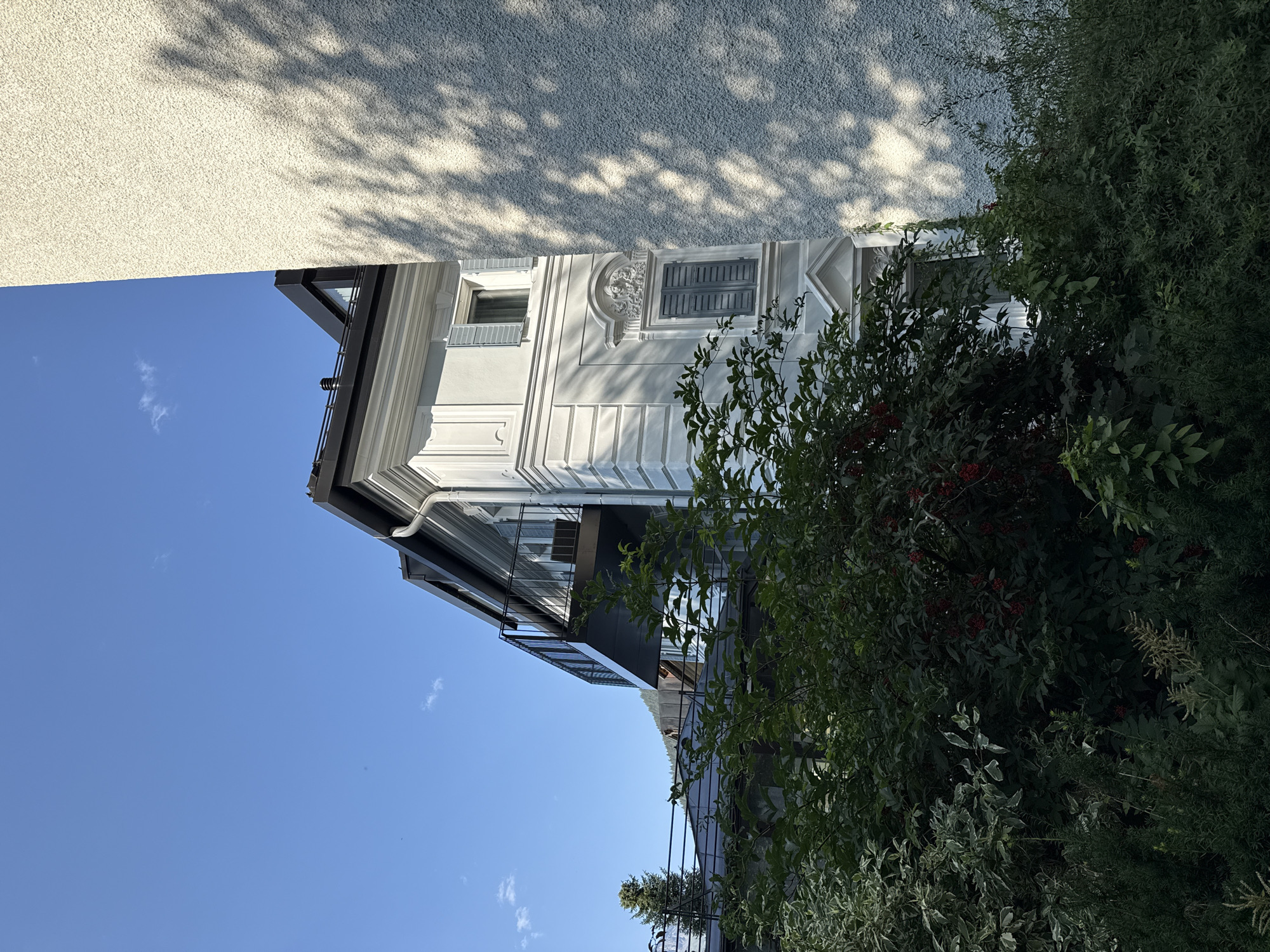 The image shows a portion of a building in Bad Gastein, Austria, with lush green foliage in the foreground. The building is modern with a white facade, black roof, and a balcony with a black railing. It features several windows, some with light blue shutters. The building is partially obscured by the dense greenery, which includes trees and bushes with small red berries. The sky is a clear, bright blue with a few small white clouds. A rough, textured wall of another building casts a shadow pattern from tree branches onto its surface, suggesting the presence of nearby trees. The scene is set during the daytime with bright sunlight.
