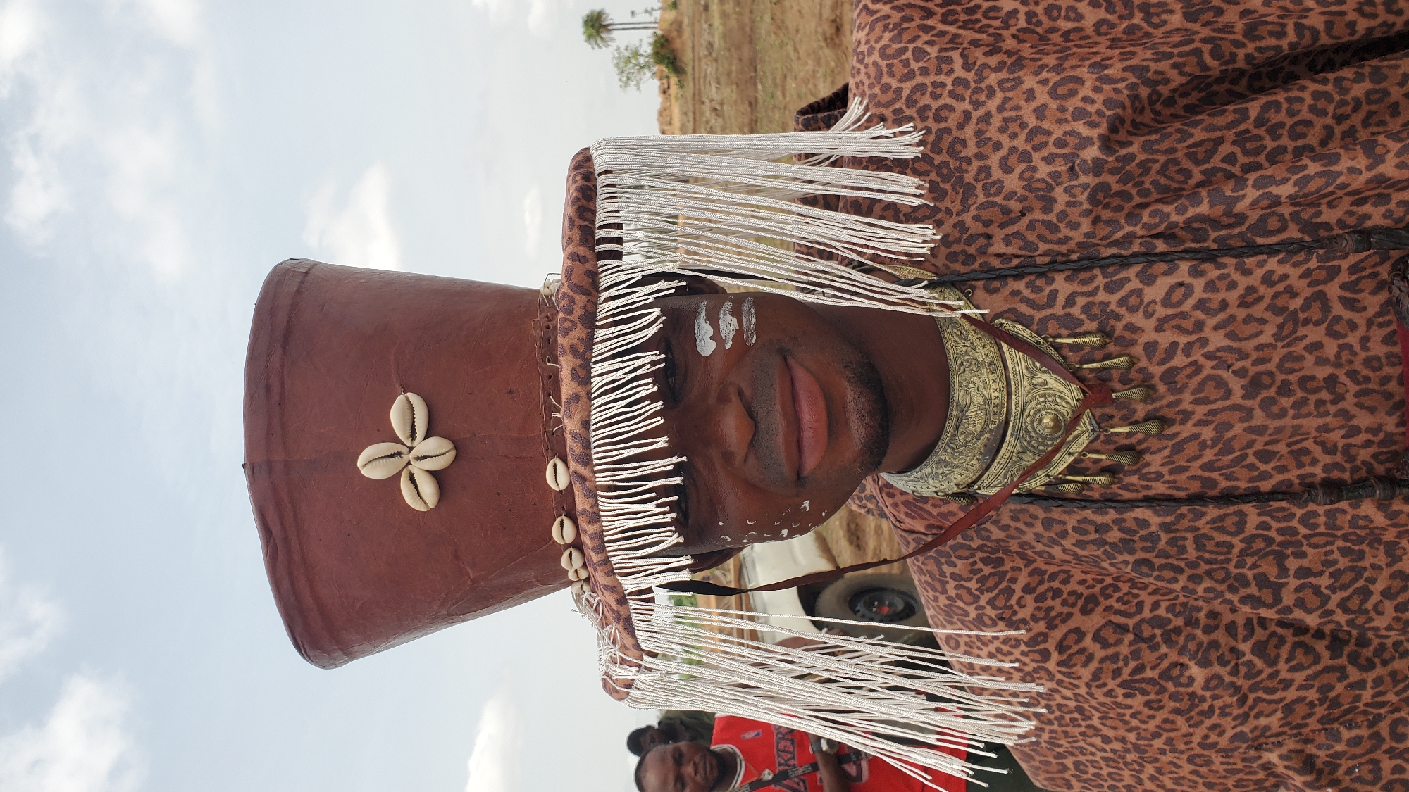 The image shows a man in traditional attire, likely in Akwanga, Nigeria. He wears a brown conical hat adorned with cowrie shells and a silver fringe. White markings are painted on his face. He also wears a leopard-print garment and an ornate gold-colored collar. The background includes a landscape with sparse vegetation and a few distant trees. Other people are visible in the lower portion of the photo. A tire is visible from a vehicle.