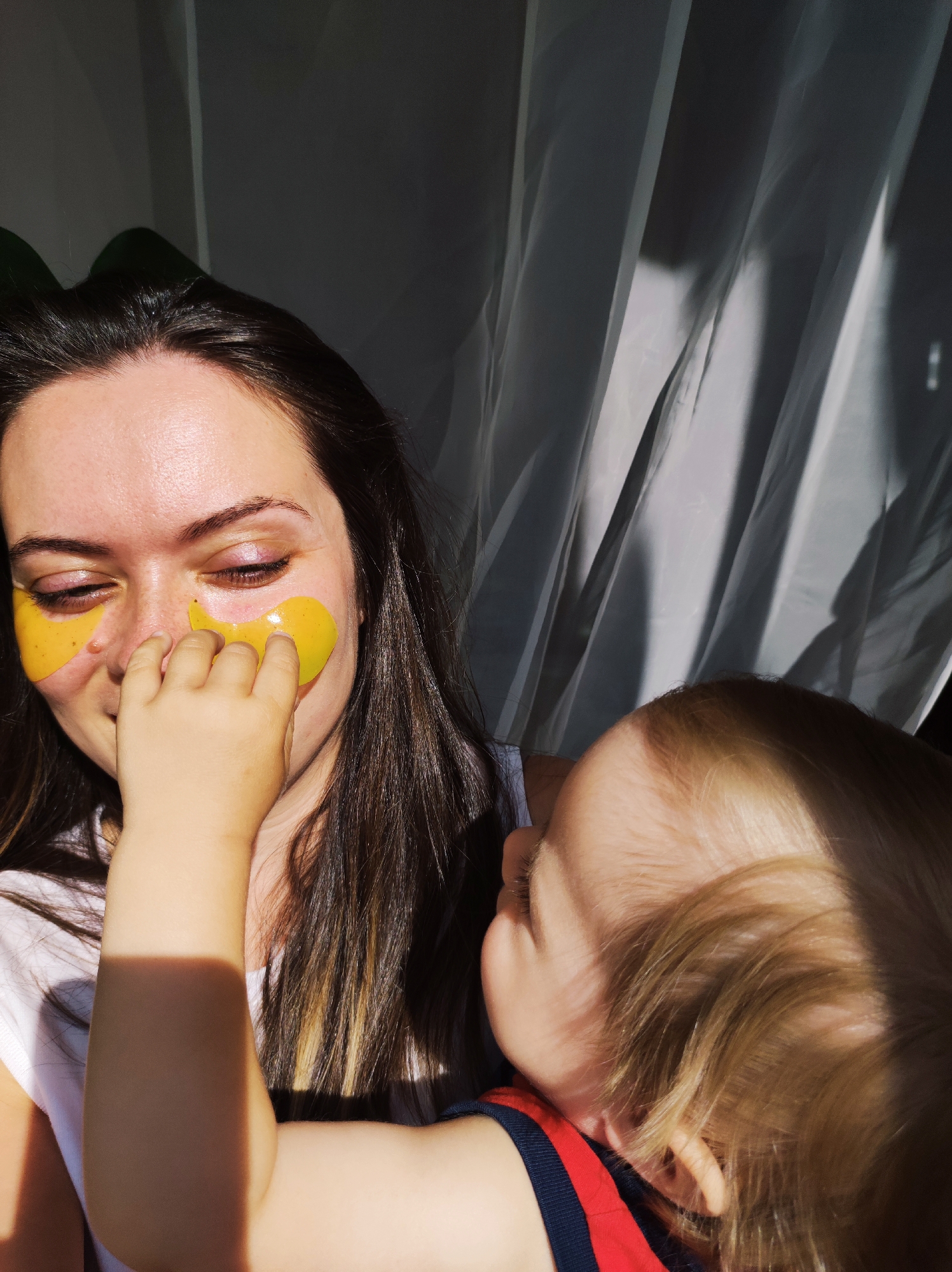 A medium shot depicts a woman with brown hair wearing yellow under-eye masks while a young child with blonde hair touches her face. The woman is smiling slightly with her eyes closed. The child is looking at the woman's face. The woman is wearing a white shirt. The child is wearing a red shirt with navy blue trim. The background consists of a white curtain and some greenery. The location is potentially a home in Chaikovsky, Russia, but there is no specific evidence in the image to confirm this.