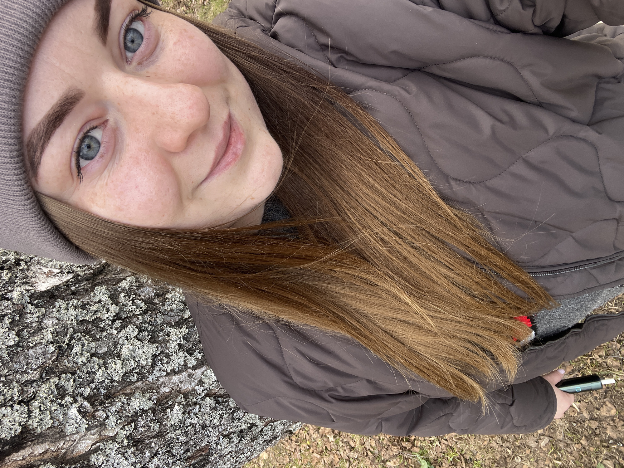 A young woman with blue eyes and light brown hair, wearing a grey beanie and a brown quilted jacket, poses for a selfie. The background shows a tree trunk covered with lichen and some fallen leaves, suggesting an outdoor location, possibly a park or wooded area. The woman is holding an electronic device in her right hand. The location is identified as Vysokovsk, Russia.