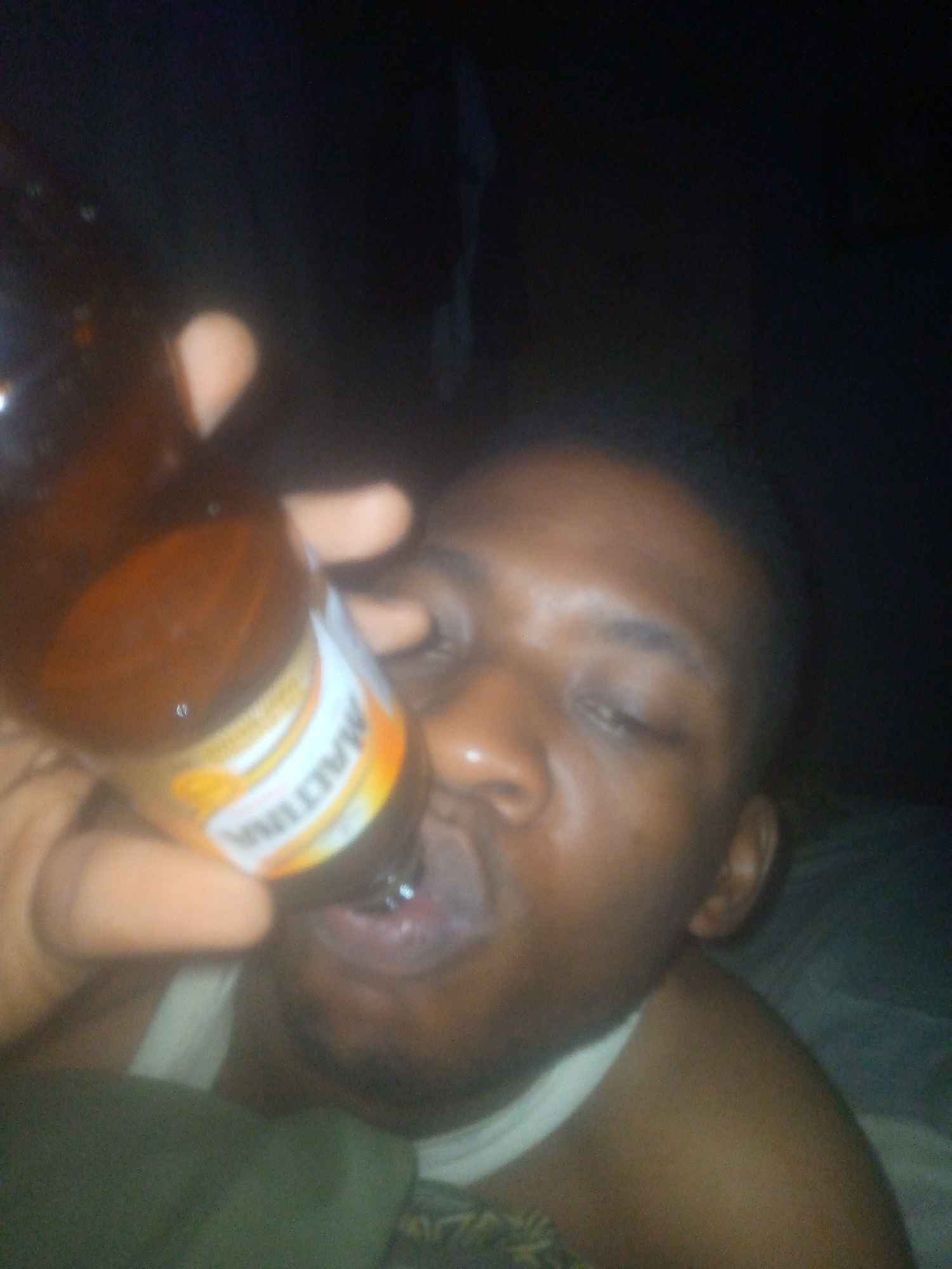 A close-up, poorly lit, somewhat blurry photo depicts a dark-skinned person drinking from a brown bottle labeled "Maltina." The person is lying down, possibly in bed, as a white tank top and bed linens are visible. The location is Unknown, Unknown.