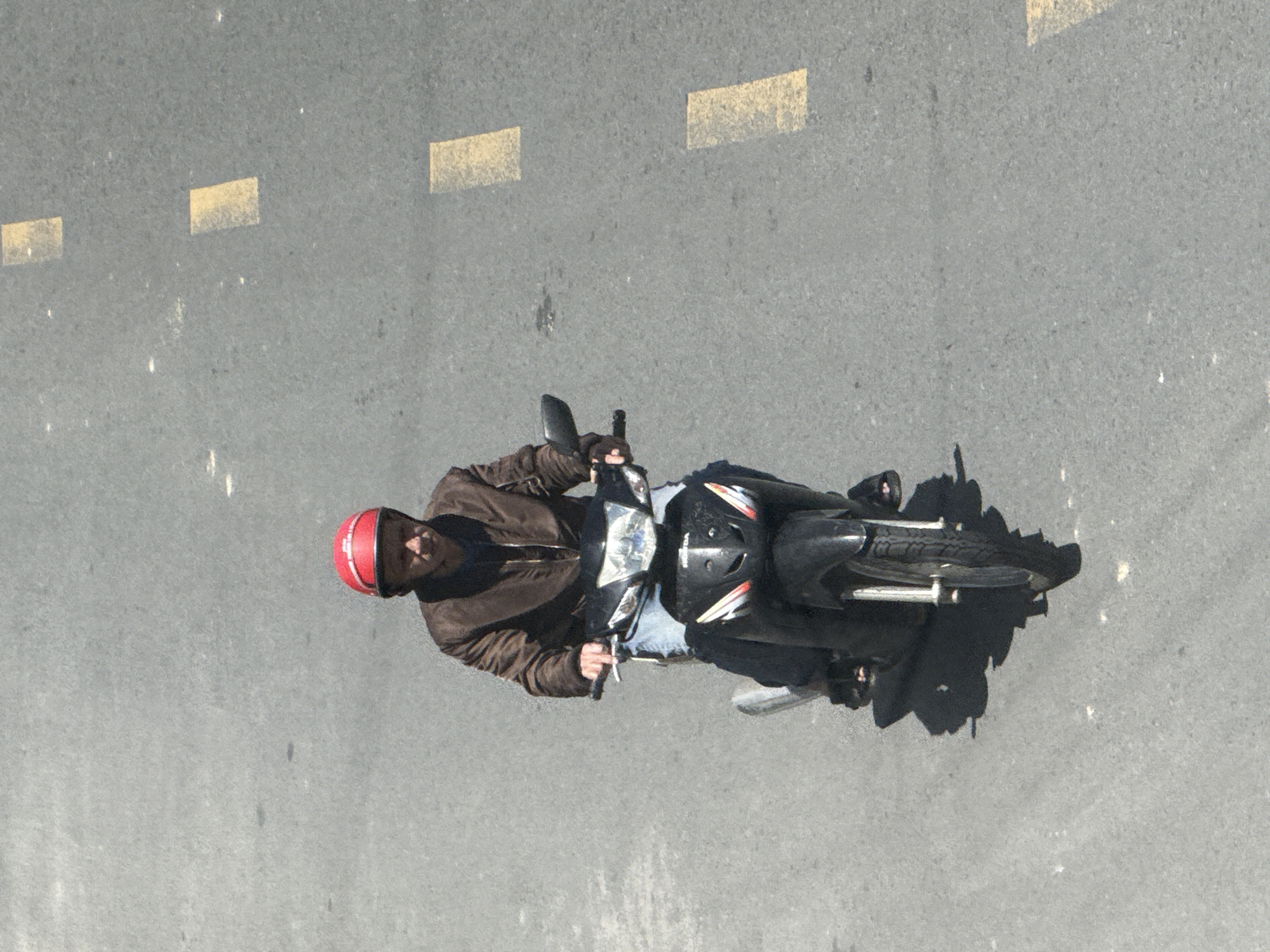 An adult male wearing a red helmet and a brown jacket rides a black scooter on a grey asphalt road. The man is looking forward, and his hands are on the handlebars. The scooter has a headlight and is partially obscured by the rider's legs. Yellow rectangular markings are visible on the road, indicating lanes or a crosswalk. The scene appears to be in an urban setting, likely Da Lat, Vietnam, given the type of scooter and road markings. The image is taken from a high angle, looking down at the scooter and rider. The lighting suggests it is daytime.