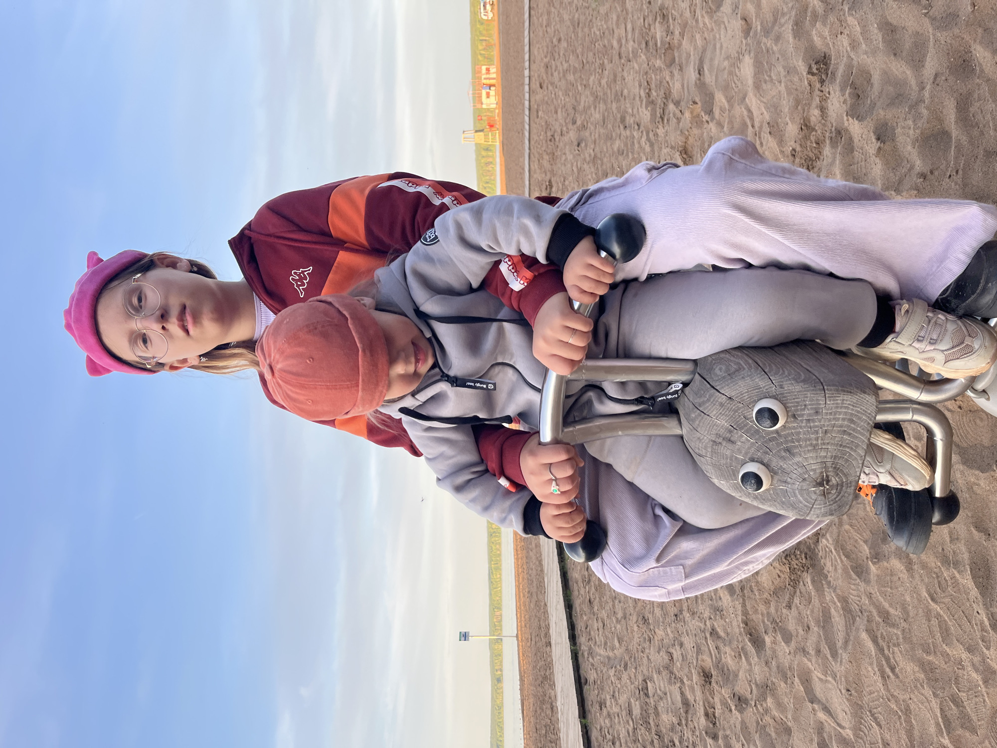 This media file, rotated clockwise, depicts two individuals, likely an older child or young adult and a younger child, on a wooden spring rider at a sandy outdoor location in Chaikovsky, Russia.
The older individual is positioned behind, wearing a pink beret with small ear-like protrusions, round-rimmed glasses, and a maroon and orange jacket featuring a white Kappa logo. Their expression is neutral. The younger child is seated in front, holding the metal handles of the spring rider. They wear a rust-colored beanie and a grey hoodie with maroon sleeves, which has a visible "® Bingoboo!" logo. Both individuals are wearing light-colored pants.
The spring rider itself is carved from wood, resembling an abstract creature or bug with large, round googly eyes. They are situated on light-colored sand, indicating a beach or playground setting.
In the background, a wide body of water, presumably the Kama River or a local reservoir, spans the mid-ground. Beyond the water, a distant shoreline with green vegetation and some industrial or urban structures is visible under a clear, pale blue sky. A single light pole with an unreadable blue sign stands near the water's edge. The scene unfolds during clear daytime weather, suggesting a peaceful and relaxed atmosphere.
