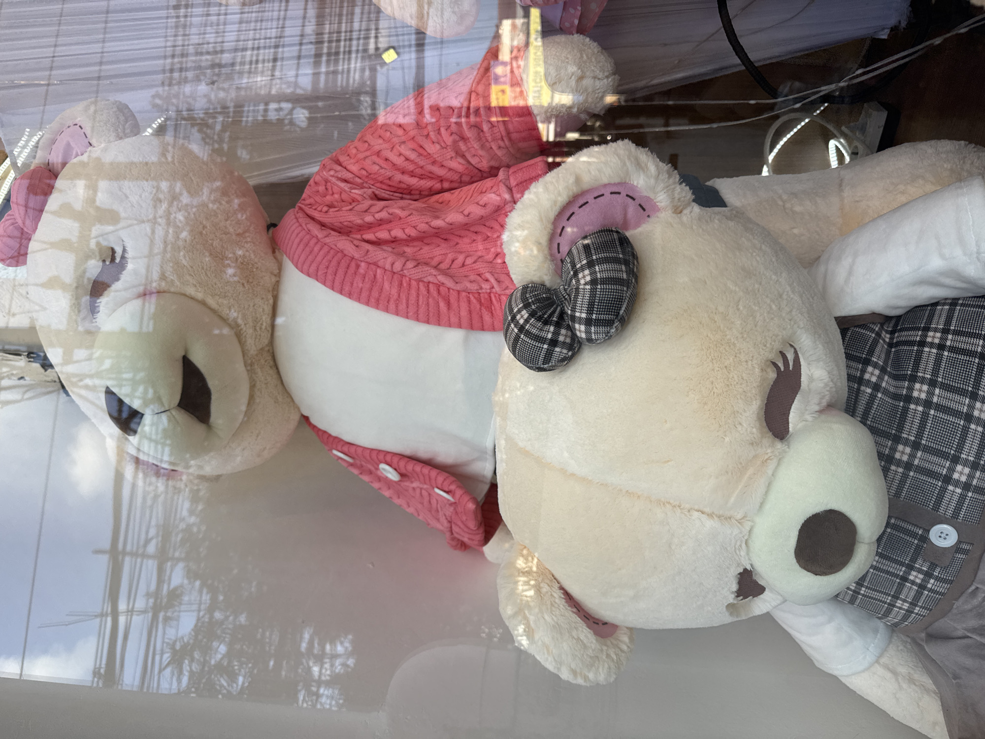 The image shows two large teddy bears on display, possibly in a shop window. The teddy bear in the foreground is cream-colored with a plaid bow on its head and a white shirt. It is positioned in front of a larger, white teddy bear wearing a pink knitted cardigan. The background is blurry and reflects the outside environment, suggesting the display is behind glass. The reflection shows trees and overhead wires, indicative of an urban setting. The lighting suggests it is daytime. There are no people visible in the image. The scene appears to be located in Denpasar, Indonesia, as indicated by the overall style and typical shop window displays found in the city.
