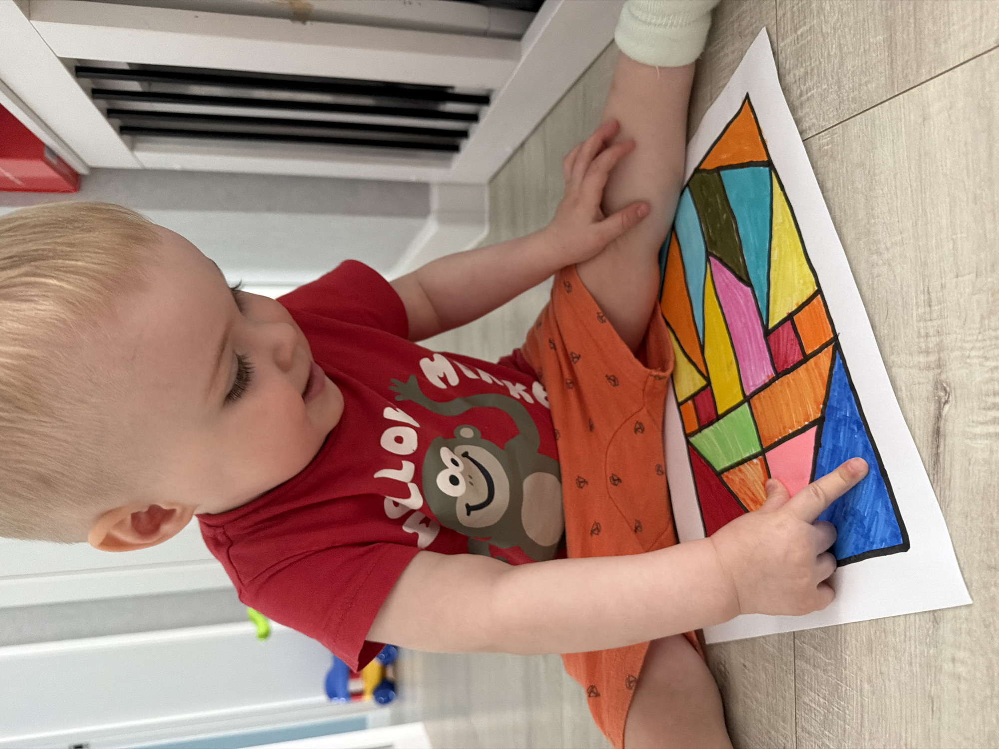 A toddler wearing a red t-shirt with a monkey graphic and orange shorts is sitting on a light-colored, wood-look floor. The child is holding a white piece of paper with a geometric drawing filled with various colors and is pointing at a blue section of the drawing. The toddler has blonde hair and is looking at the drawing with a slight smile. Part of a white cabinet or furniture piece with horizontal slots is visible in the background. Multi-colored toys are partially visible in the lower left corner of the image. There is no information to suggest the photo was taken in Votkinsk, Russia.