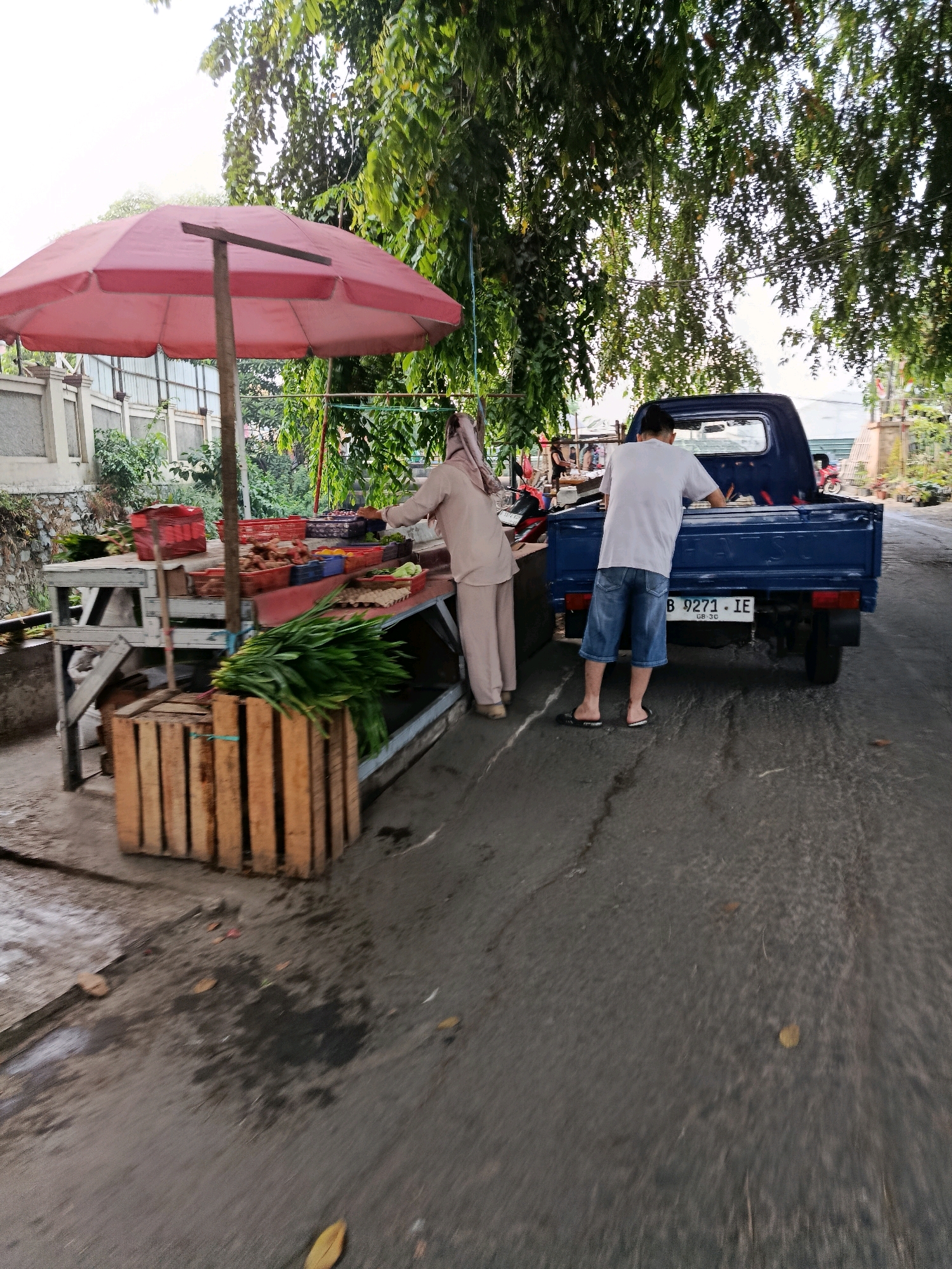 A man in a white t-shirt and blue denim shorts stands near the back of a blue pickup truck, facing it. A woman in a beige hijab and matching outfit stands behind a market stall to the left, interacting with the produce. The market stall is laden with various fruits and vegetables, with a red umbrella providing shade. Wooden crates are stacked beside the stall. The scene appears to be on a paved road, possibly in an outdoor market setting, with lush green trees overhead. The license plate of the truck reads "B 9271 IE." The overall atmosphere suggests a typical day at a roadside market in Bekasi, Indonesia, with a focus on commerce and everyday life.