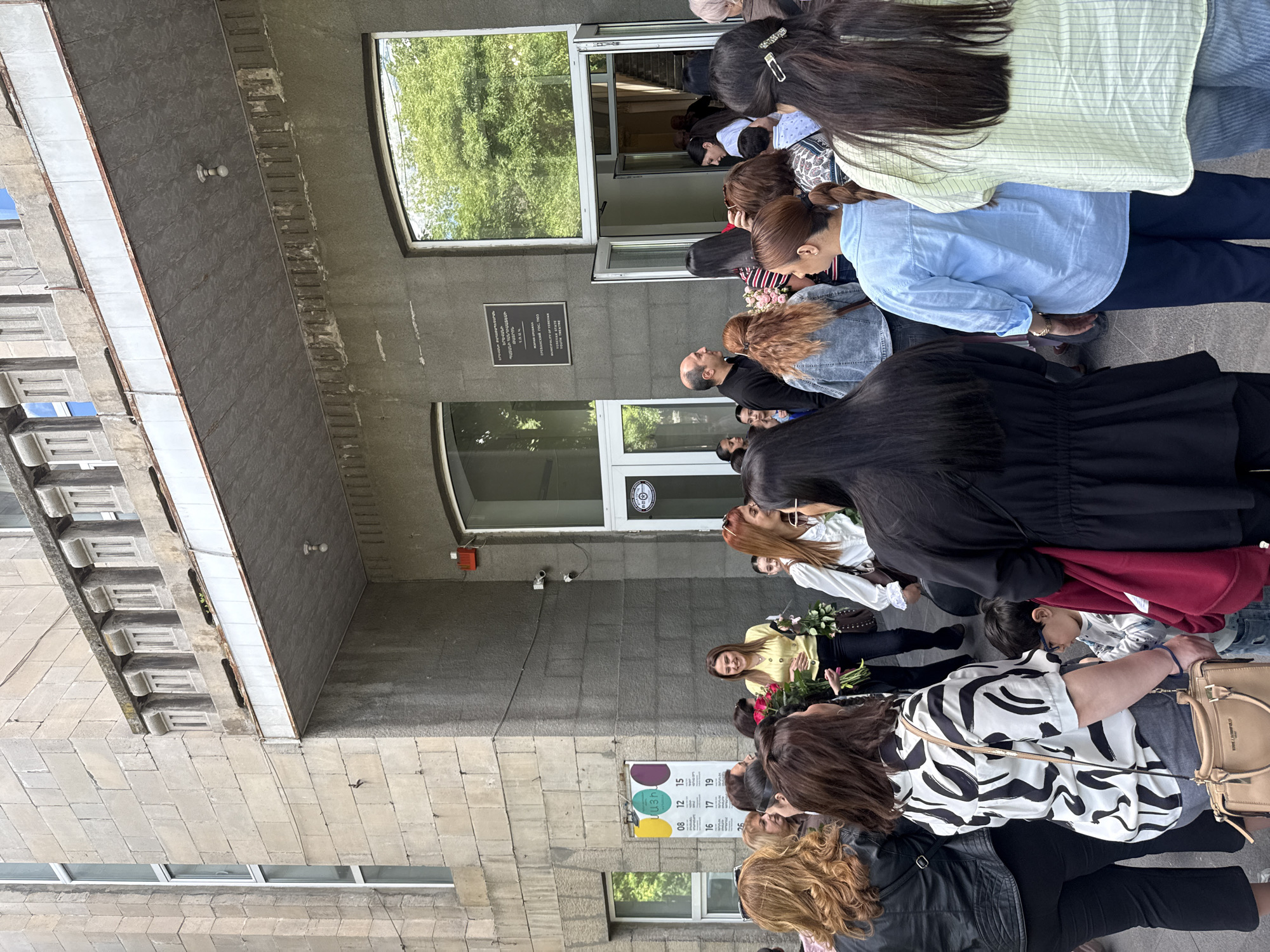 A ground-level shot depicts a crowd of people gathered outside a gray stone building. Several individuals are carrying bouquets of flowers. A sign with numbers is visible on the building's exterior. A plaque with text written in both Armenian and English is visible above a doorway. Based on the Armenian text on the plaque and the building's architectural style, the location is likely Yerevan, Armenia.