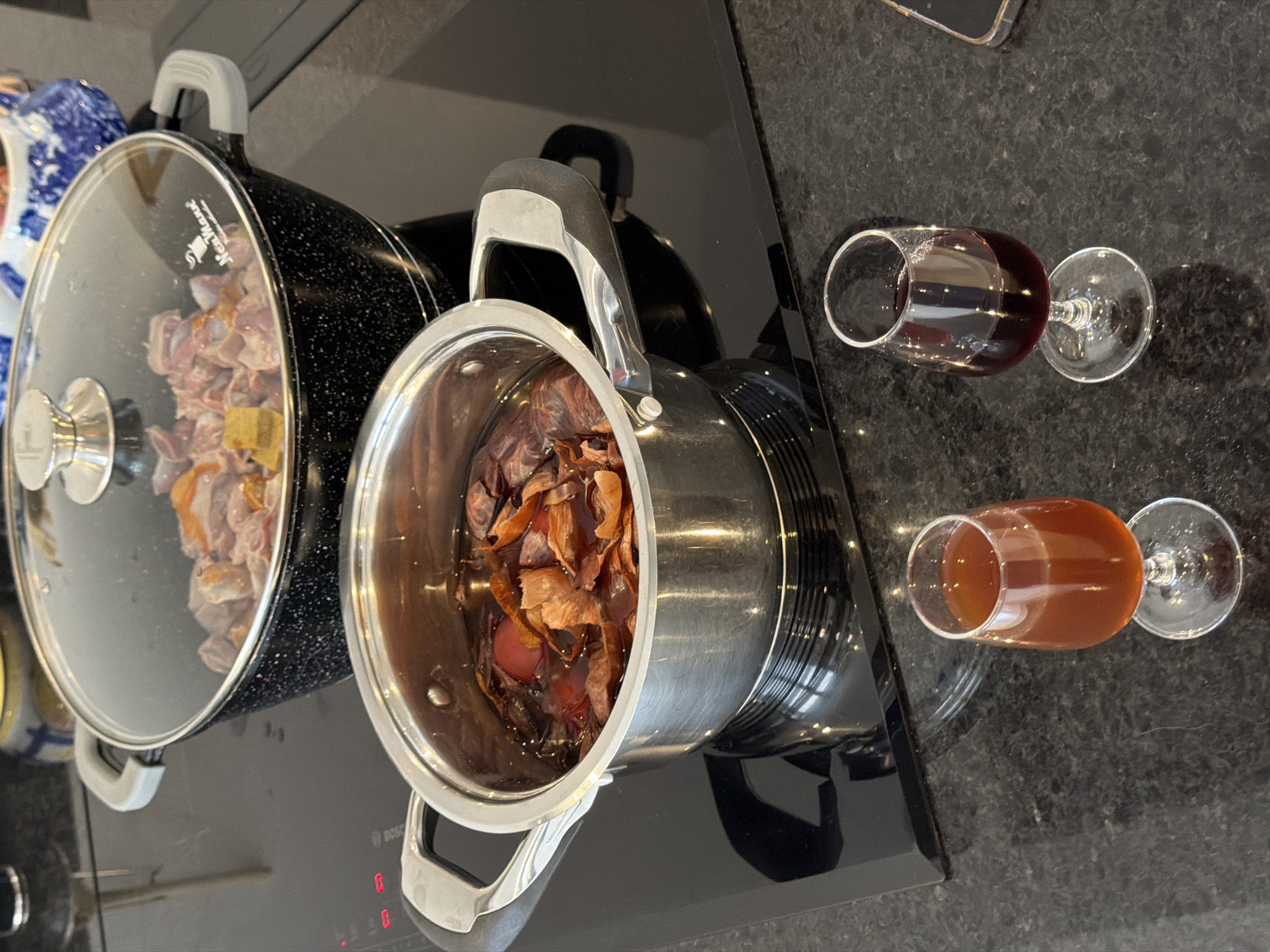 Two pots sit on a black cooktop, one containing meat, the other, onion skins. Two wine glasses are nearby, one filled with red wine, the other with a lighter-colored beverage. The location appears to be a kitchen, based on the cooktop and countertop. There are no people visible in the image. The scene is likely indoors, with natural lighting. The image was taken in Esch-sur-Alzette, Luxembourg.