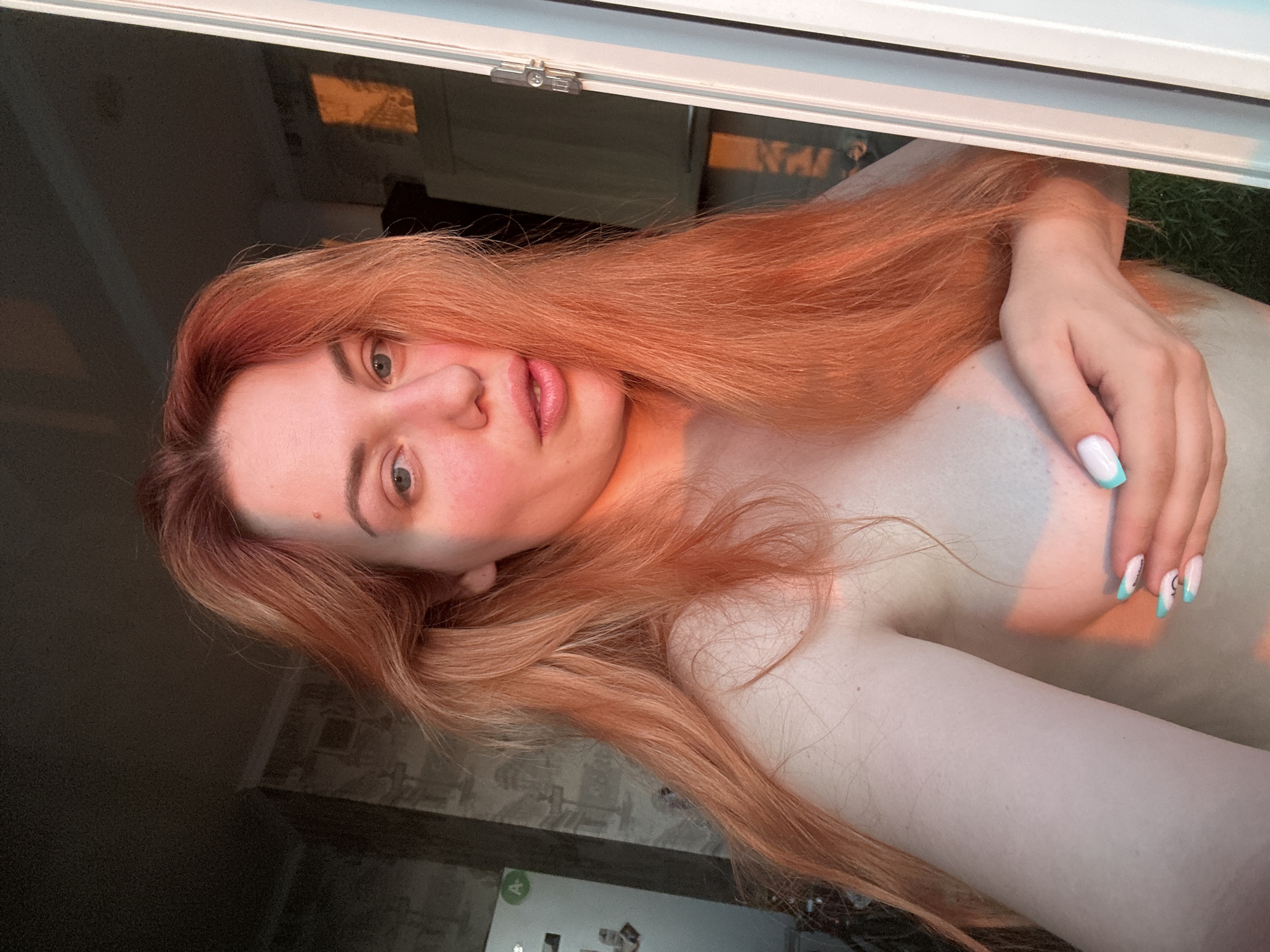 A young woman with long, reddish-orange hair is shown from the chest up, holding her left hand over her chest. She is facing the camera, with her head tilted slightly. Her eyes are open and looking towards the camera. The lighting suggests it is either sunrise or sunset, casting a warm, orange glow on her skin and hair. She is indoors, with a window frame visible to her right. The background is blurred, but appears to be a room with wallpaper and a door.