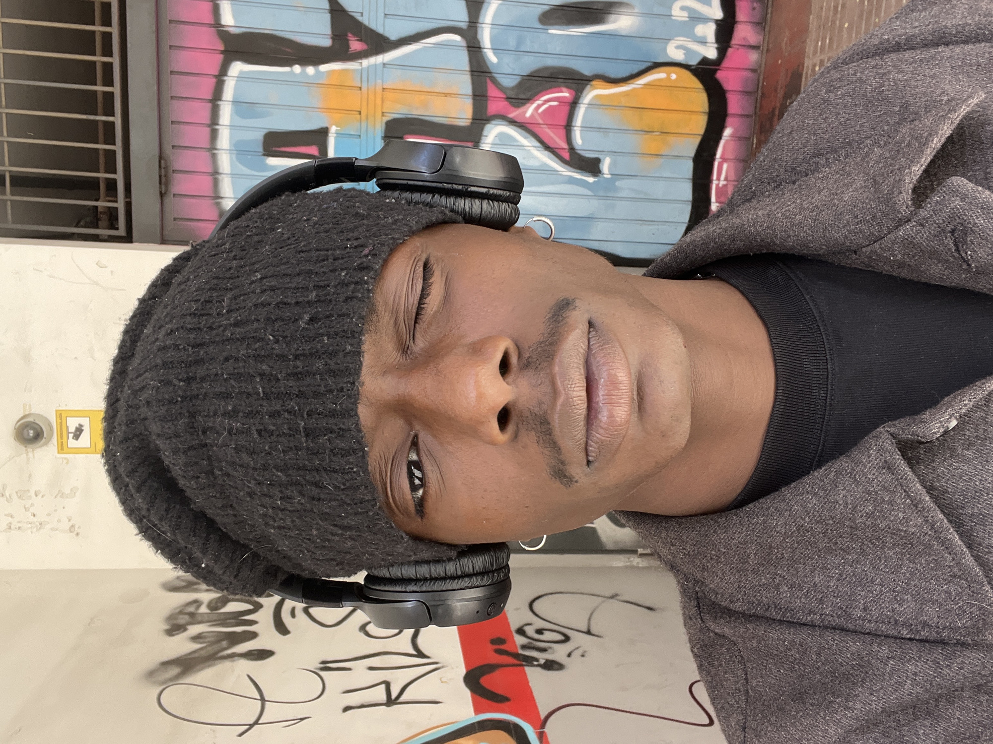 A medium shot presents a person of African descent. They are wearing a dark-colored beanie and over-ear headphones, and a gray tweed coat. The subject’s right eye is slightly closed, giving a pensive or contemplative look. The person has a prominent mustache and wears a small hoop earring.
The backdrop is graffiti-covered walls, indicative of an urban environment. The graffiti includes colorful shapes and text, suggesting a street art scene. A window with metal bars is visible in the background, along with other elements of urban infrastructure like a security camera.
While the scene is urban, there's no definitive information to confirm the specific location in Barcelona. However, the presence of graffiti and a window with metal bars is typical of certain urban areas in Barcelona and other European cities.