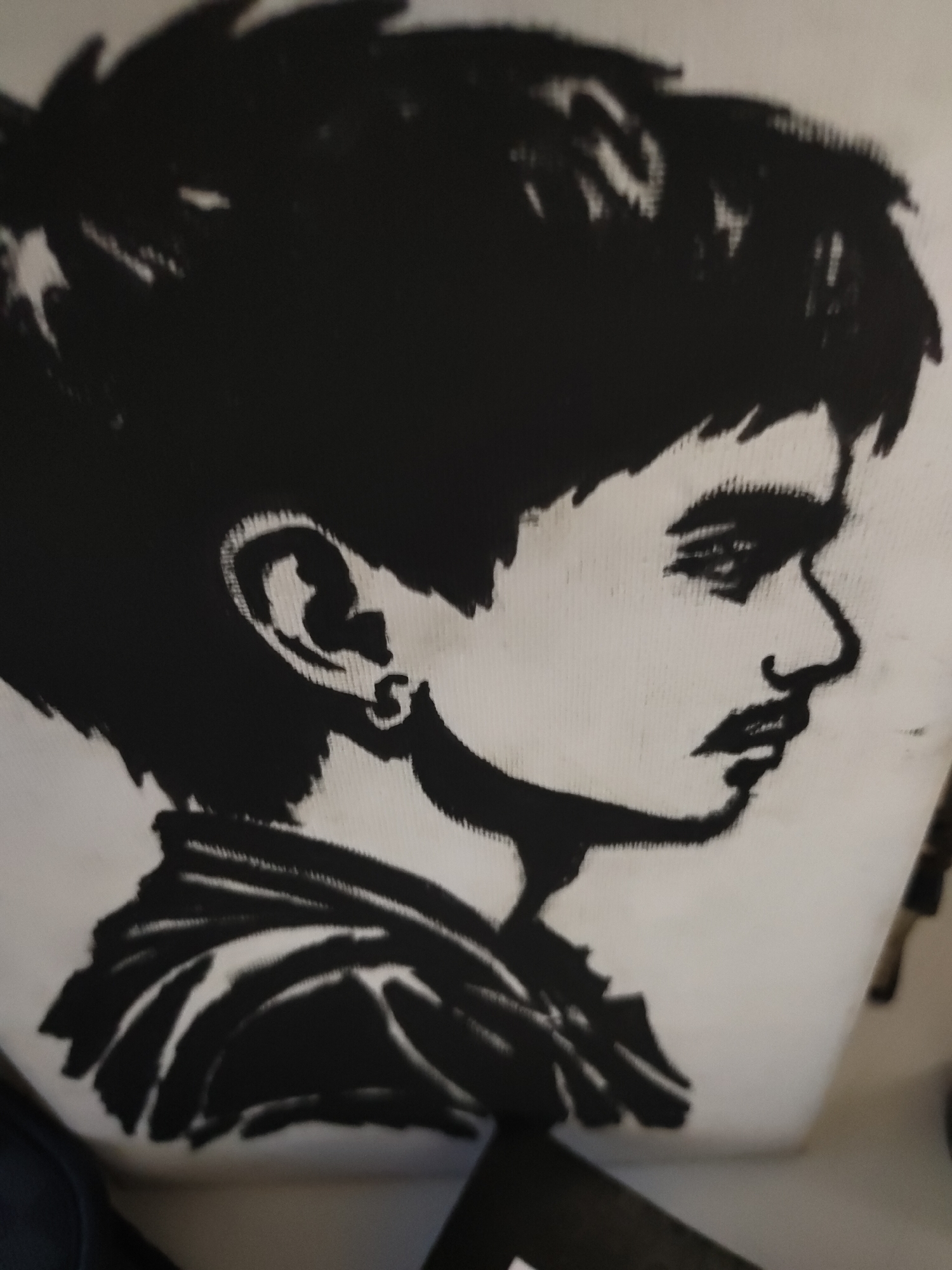 The image displays a close-up of a monochrome graphic print, rendered in solid black against a light-colored, possibly off-white, textile surface, indicative of clothing or a canvas item. The print depicts a stylized profile portrait of a young person facing right. The individual features voluminous, spiky black hair that rises from the head and extends backward. The face shows a distinct brow, a defined nose, and lips that are slightly parted, with a subtle mustache visible above the upper lip. The visible ear contains a small, circular earring in the lobe. The neck is short, leading to broad shoulders covered in clothing, depicted with multiple dark folds and lines. The background surrounding the printed object is blurred and indistinct, preventing identification of any specific objects, structures, or an overall environment. The precise location, including the city of Davao City, Philippines, cannot be determined from the visual evidence within the image.
