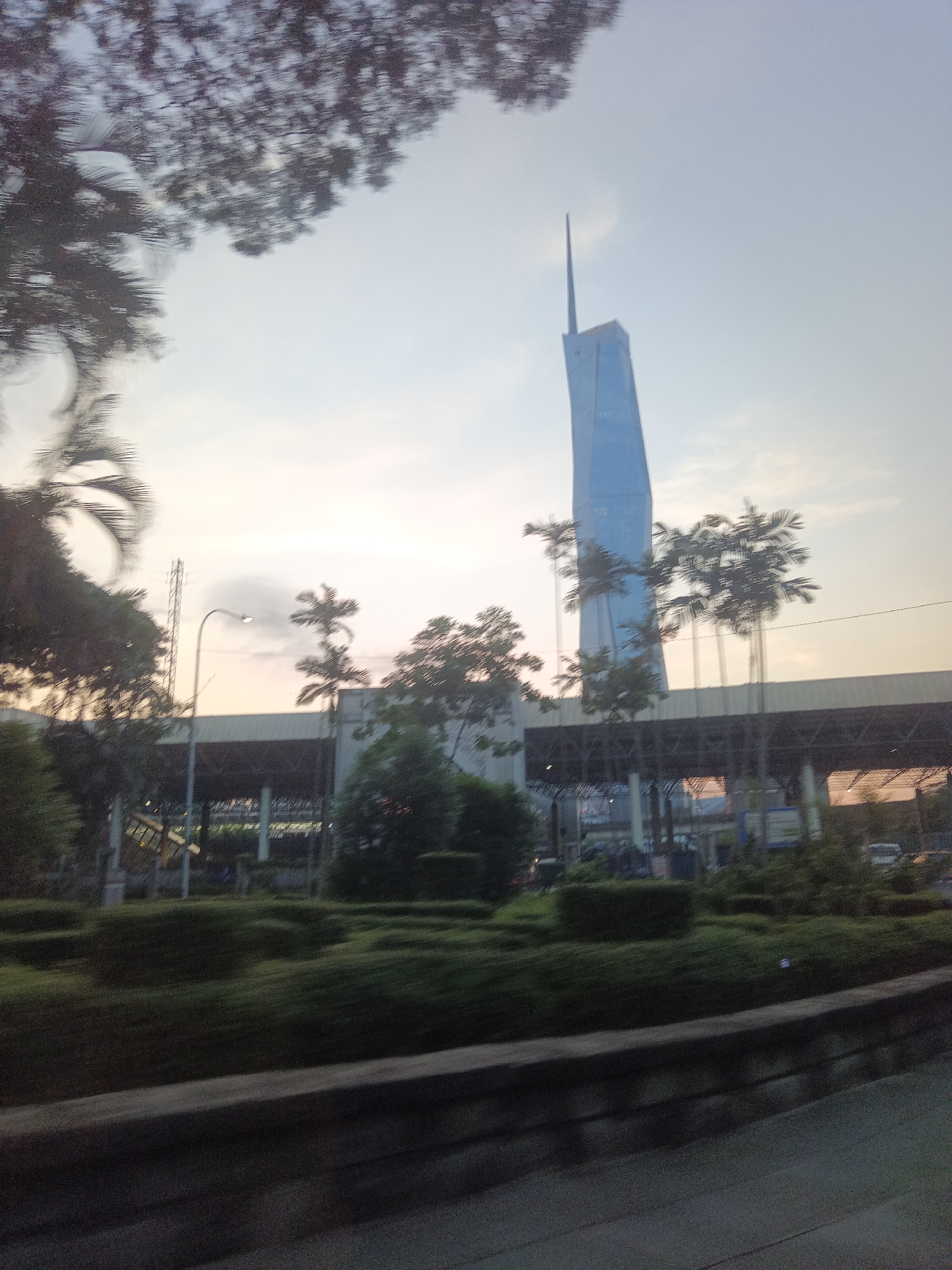 The image depicts a scene in Kuala Lumpur, Malaysia, featuring the distinctive Merdeka 118 tower, the second-tallest building in the world, under a hazy sky. The tower, a light blue skyscraper with a pointed spire, dominates the upper right portion of the frame. In the foreground, lush green foliage and manicured hedges are visible, suggesting a well-maintained park or roadside landscaping. A dark grey concrete barrier runs along the bottom of the image. Behind the greenery, a building with a dark, covered structure and supporting beams is partially obscured by trees and palm trees. A streetlamp with a single light is positioned centrally, casting a faint glow. The overall impression is one of a city view, likely captured during twilight or early morning, with hints of movement blurring some elements, suggesting the photo was taken from a moving vehicle.