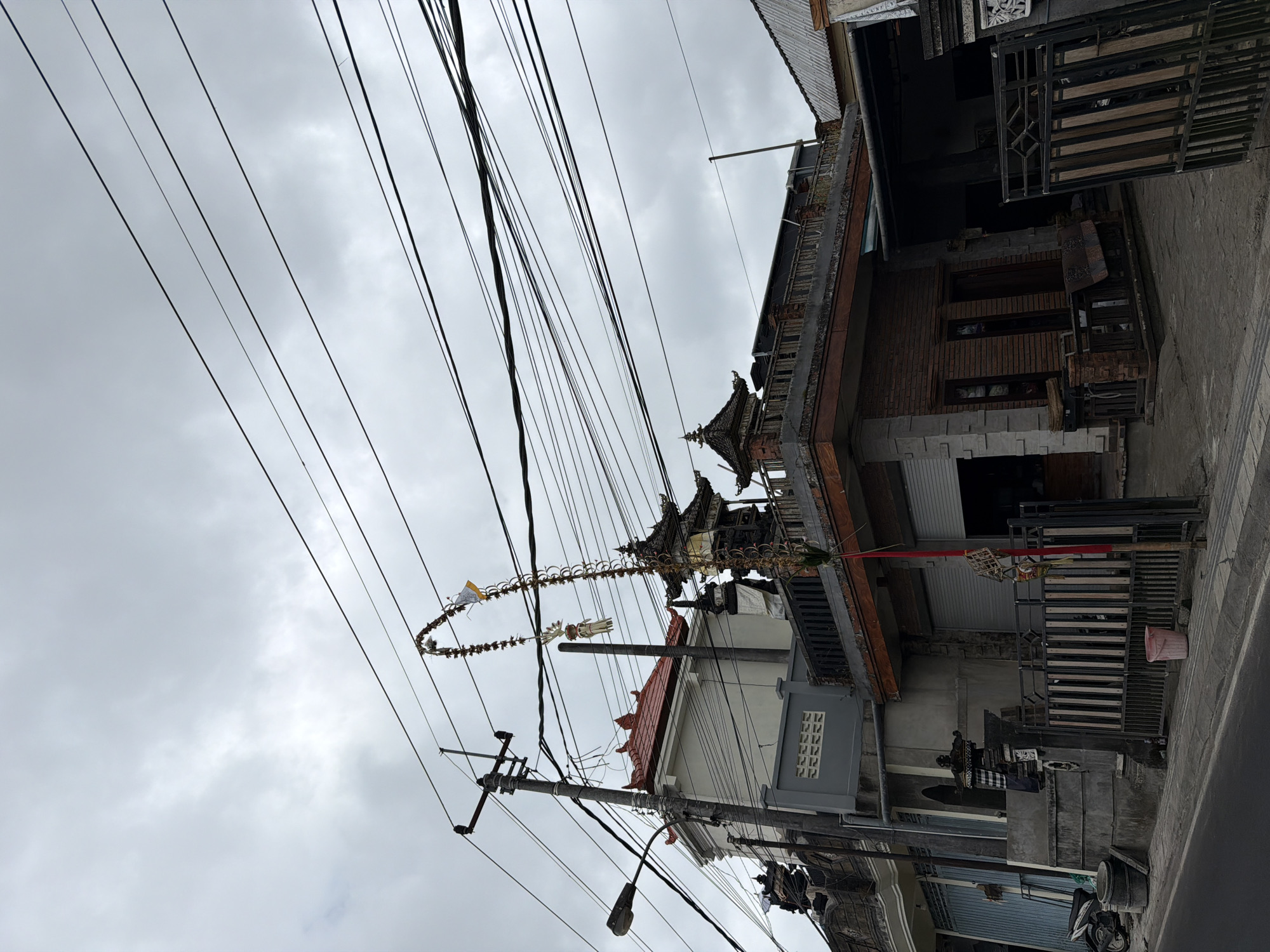 The image depicts a street scene in Kintamani, Indonesia, characterized by a cloudy sky overhead. Dominating the composition are numerous black electrical wires crisscrossing the frame, suspended from a utility pole on the left. Below the wires, a multi-story building with a red-tiled roof and grey siding is visible, alongside a brick building with a dark wooden balcony and a corrugated metal roof to the right. A decorative, curved structure, possibly an offering or a traditional decoration, hangs from the utility pole and drapes over some of the wires. The ground level features a paved sidewalk, a street, a metal fence, and various objects including a red bucket, stacks of what appear to be concrete drums, and a motorcycle. Architectural elements of the buildings suggest a Balinese influence, with tiered roofs and ornate details on some structures.