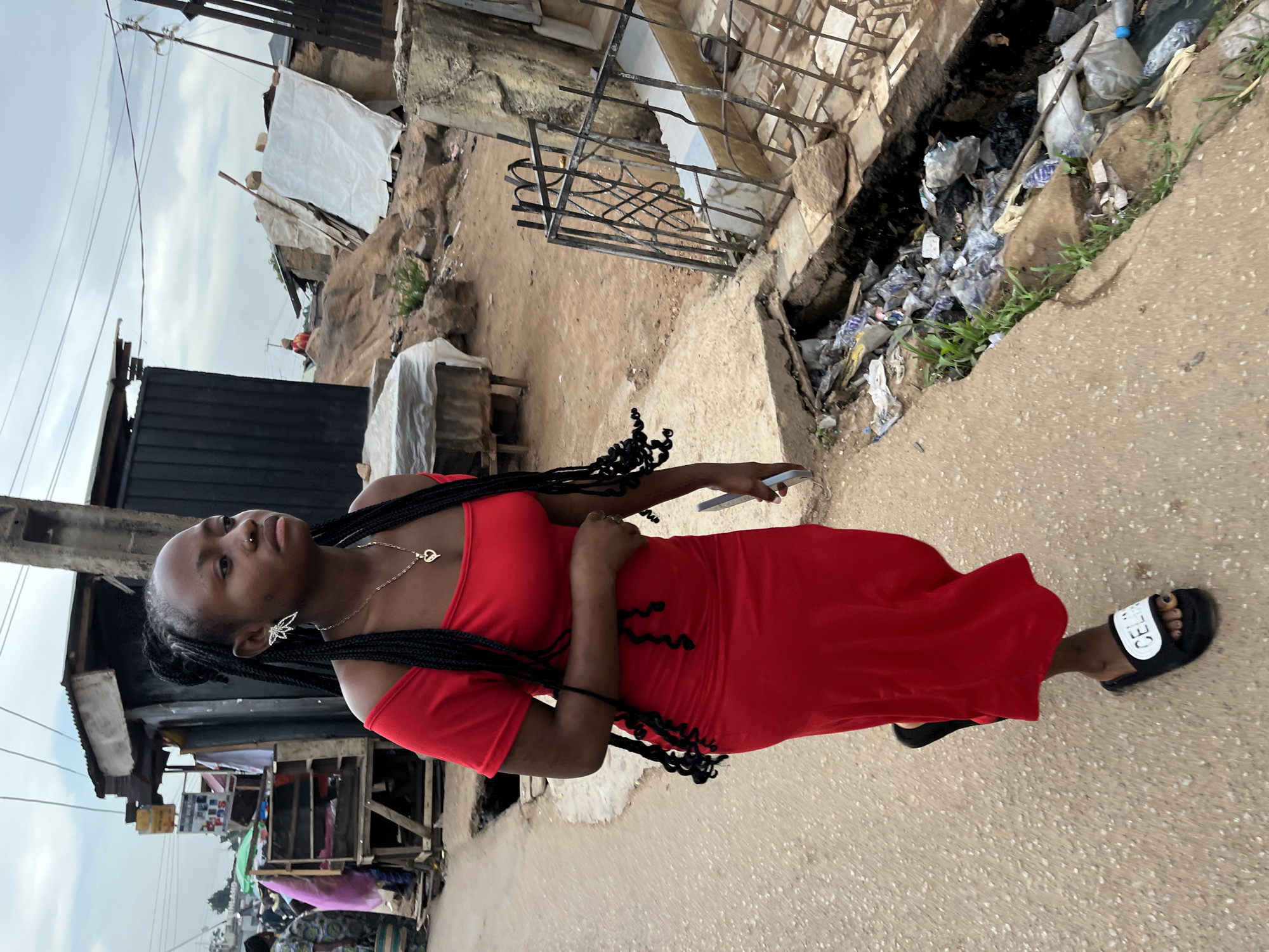 A young Black woman in a red dress stands on a concrete sidewalk in Jos, Nigeria. She faces left, looking up and away from the camera, and holds a phone in her right hand. Her hair is styled in long, dark braids. She wears a gold necklace and dangling earrings. To the left, there are makeshift buildings and shops constructed with corrugated metal and wood, suggesting a market area. In the background, the environment appears urban, with a mix of structures. To the right of the sidewalk, there is a deep ditch filled with trash and debris. The sky is overcast.
