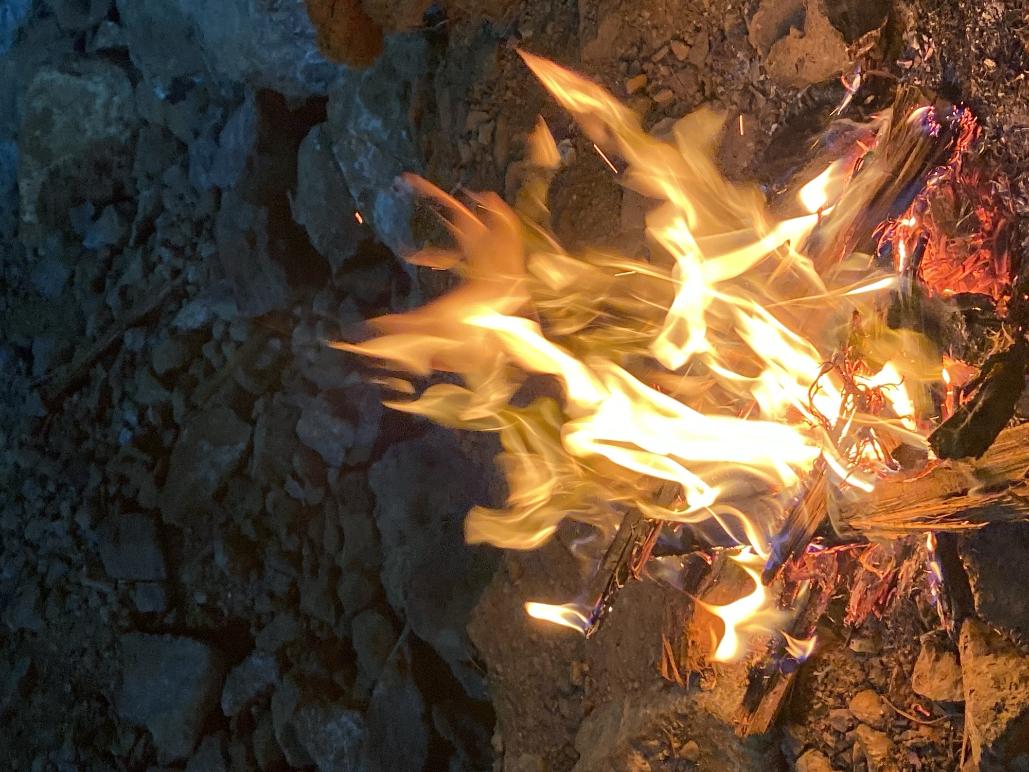 The image shows a close-up, low-angle view of a small campfire burning outdoors on rocky ground. The flames are a vibrant orange and yellow, with some red embers visible at the base of the fire. Several pieces of wood are burning, some appearing charred and others still intact. The background consists of dark, irregularly shaped rocks and dirt, suggesting a natural, possibly rugged environment. There are no people visible in the image. The scene is captured at night or in low light conditions, as indicated by the darkness of the surroundings and the illumination provided solely by the fire.
