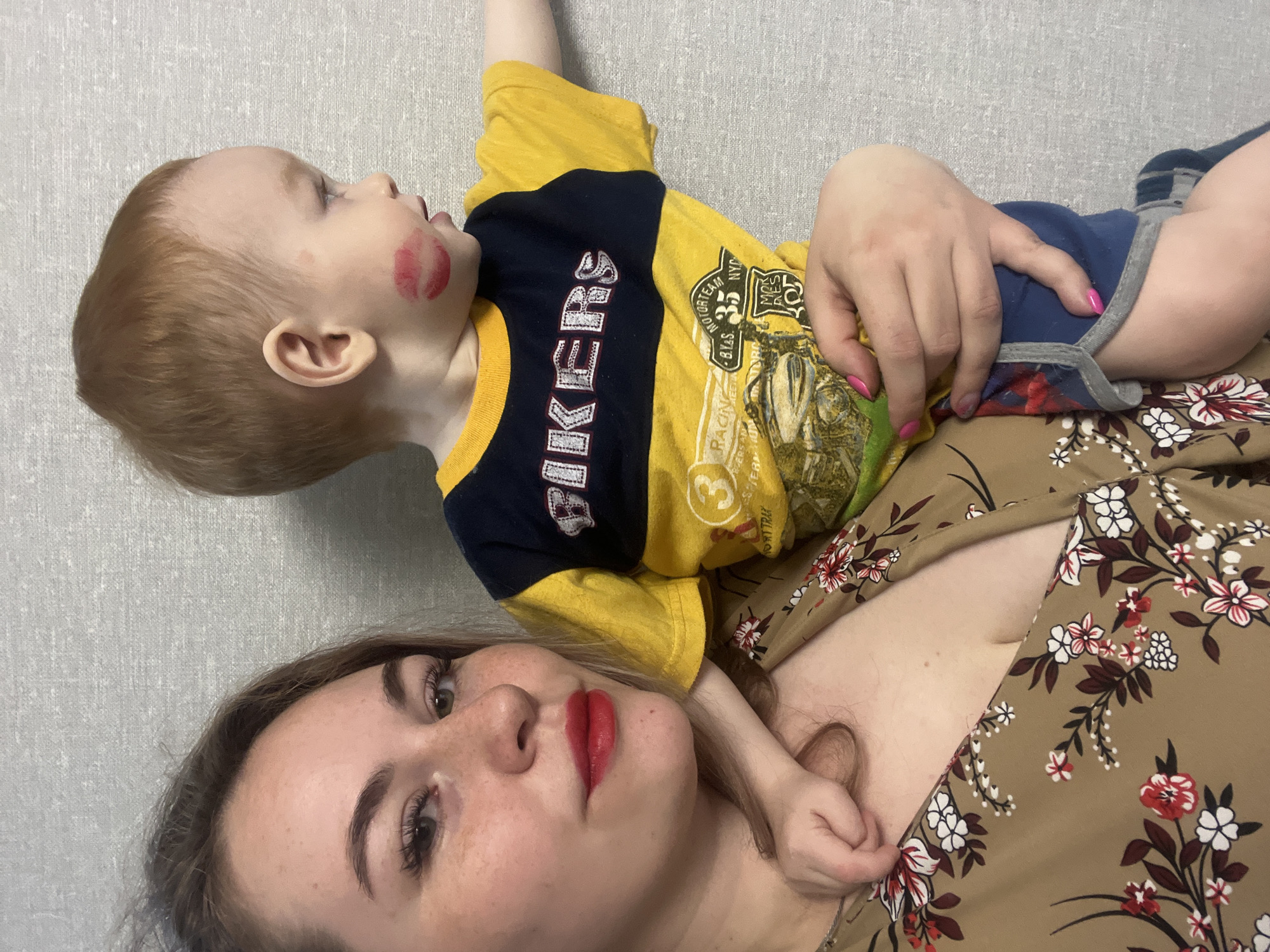 A young child with a lipstick mark on their face is lying on top of a woman. The child has red hair, and wears a yellow and navy blue shirt with an image on the front. The woman is wearing a floral-patterned brown shirt and has red lipstick on. The background appears to be a plain grey wall. There is no information present to determine the location is in Chaikovsky, Russia.