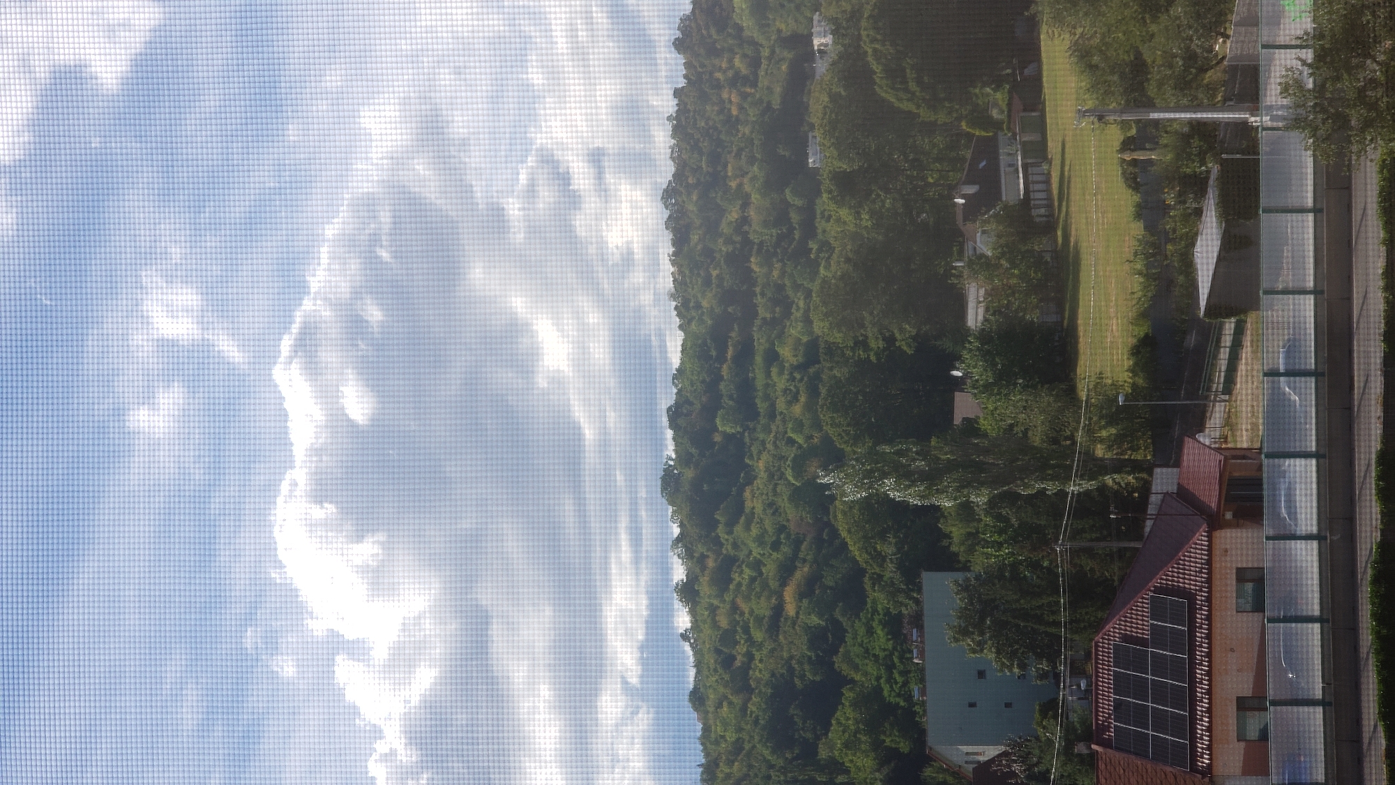 This is a photograph taken from an elevated position looking out over a suburban landscape. The foreground features a highway barrier with some foliage visible below it. Beyond the barrier, there is a red-roofed house with solar panels installed on its roof. To the left of this house is a light blue rectangular building. Further back, a hillside covered in lush green trees dominates the midground. The sky above is a mix of blue with scattered white, fluffy clouds. The image appears to be taken through a mesh screen, as evidenced by the grid pattern overlaid on the scene. The presence of solar panels and the architectural styles suggest a residential area. Given the context of Prešov, Slovakia, this scene depicts a view from a residential building or viewpoint looking towards a forested hillside and suburban elements within the city or its immediate surroundings. There are no people or animals visible in the photograph.