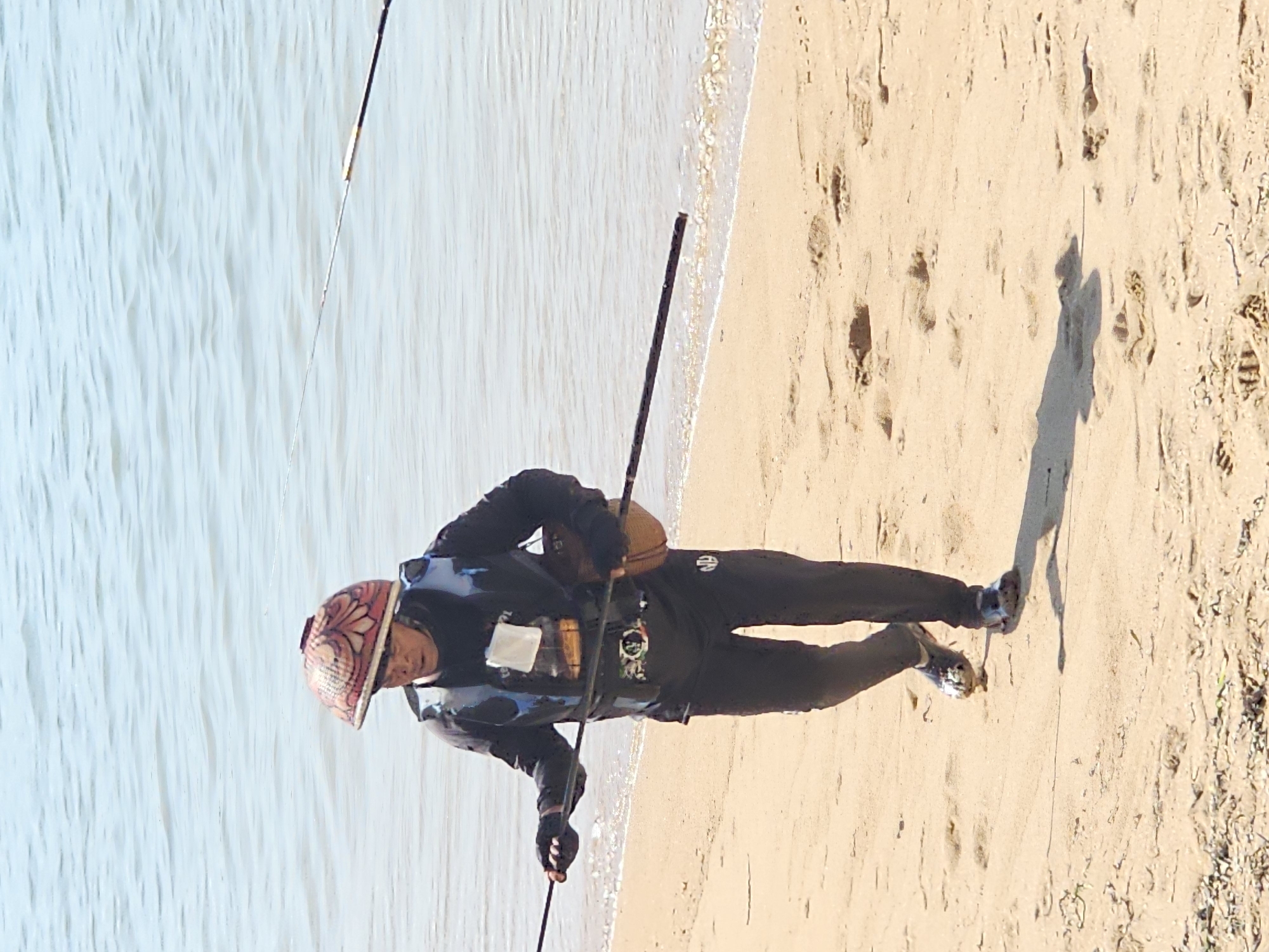 The image shows a person walking on a sandy beach with the ocean in the background. The person is wearing a dark long-sleeved shirt, dark pants, a life vest, and a conical hat. They are carrying a long pole that extends horizontally across their body, and on their right side, they are carrying a round, brown object that appears to be a basket or container. The person is walking towards the viewer, with their left foot forward. The beach is sandy with some debris scattered on it. The water is calm and light blue, with small waves lapping at the shore. The scene is consistent with a location on the coast of Denpasar, Indonesia, where such activities might occur.
