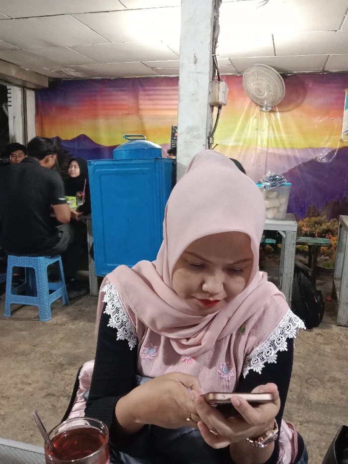 An interior scene in Kumpulrejo, Indonesia, depicts a casual gathering space. In the foreground, a woman with light skin, wearing a light pink hijab, a black long-sleeved shirt, and a pink overshirt with white lace trim and floral embroidery, is seated and looking down at a light-colored smartphone held in both hands. She has red lipstick and wears a rose gold-toned watch on her left wrist. A glass containing a dark reddish-brown liquid with a spoon is visible on a surface to her immediate left.

In the mid-ground, to the left, a man wearing a black t-shirt is seated on a light blue plastic stool, with his back partially to the camera. Behind him and further to the left, a woman in a black hijab is seated at a table, with a drink in front of her. Another individual is partially obscured behind the man in the black t-shirt. A large, rectangular blue metal container with a lid stands prominently in the mid-ground.

The background wall features a mural depicting a landscape with mountains under a multi-colored sky in hues of orange, yellow, and purple. A rough, light-colored concrete pillar is visible on the right side of the image, with electrical wiring and a small wooden box attached. A white wall-mounted oscillating fan is affixed to the right wall above the mural. On a table to the right, behind the foreground subject, are plastic containers holding various items, possibly food. The ceiling is white and paneled, with visible light sources. The floor appears to be concrete or unpaved earth.