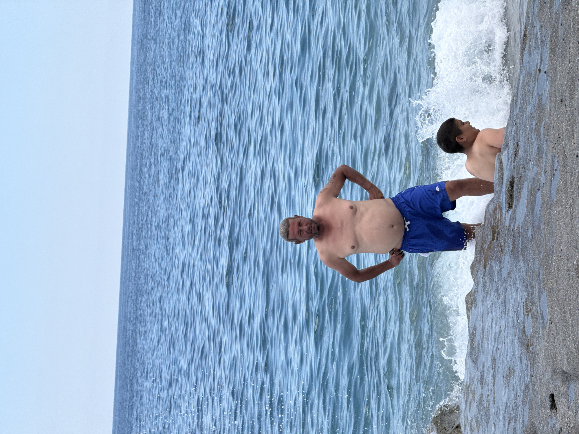 A medium, angled shot depicts a man and a child at the edge of a body of water. The man, older with graying hair, is shirtless and wears blue swim trunks. He stands facing the water with his hands on his hips, appearing to look directly ahead. To his right, a child with dark hair and bare back is partially obscured by a wave of water. They are positioned at the edge of a rocky shoreline. The water stretches out towards the horizon under a pale sky. The location is Unknown, Unknown.
