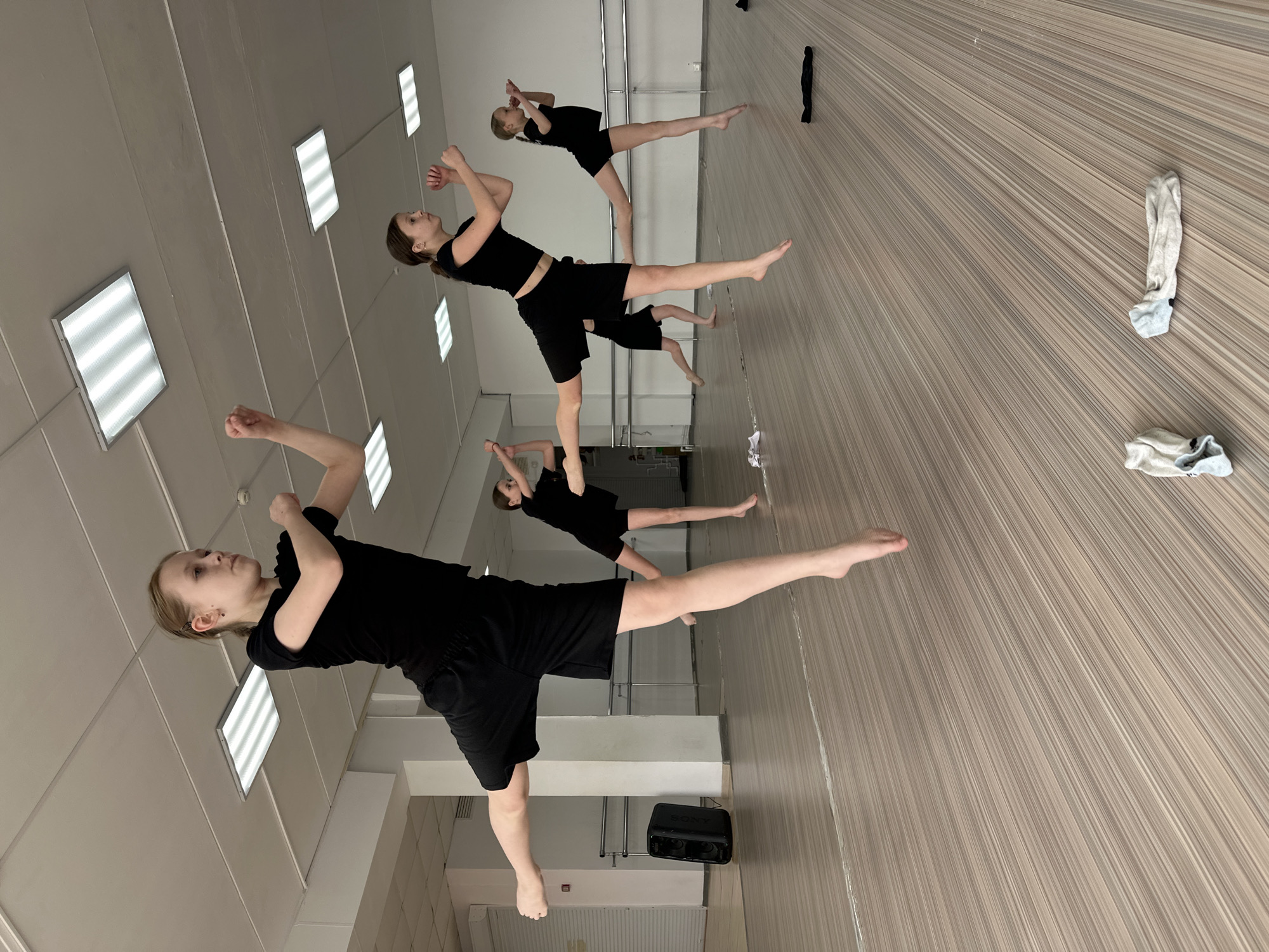 In a dance studio in Votkinsk, Russia, a group of five girls practice a dance routine. They are all dressed in black crop tops and shorts. The girls are in various poses, some with arms raised and legs extended. The studio has a light wood-grain floor and white walls with rectangular fluorescent light fixtures. A black Sony speaker is on the floor near one of the girls. There are scattered socks on the floor.