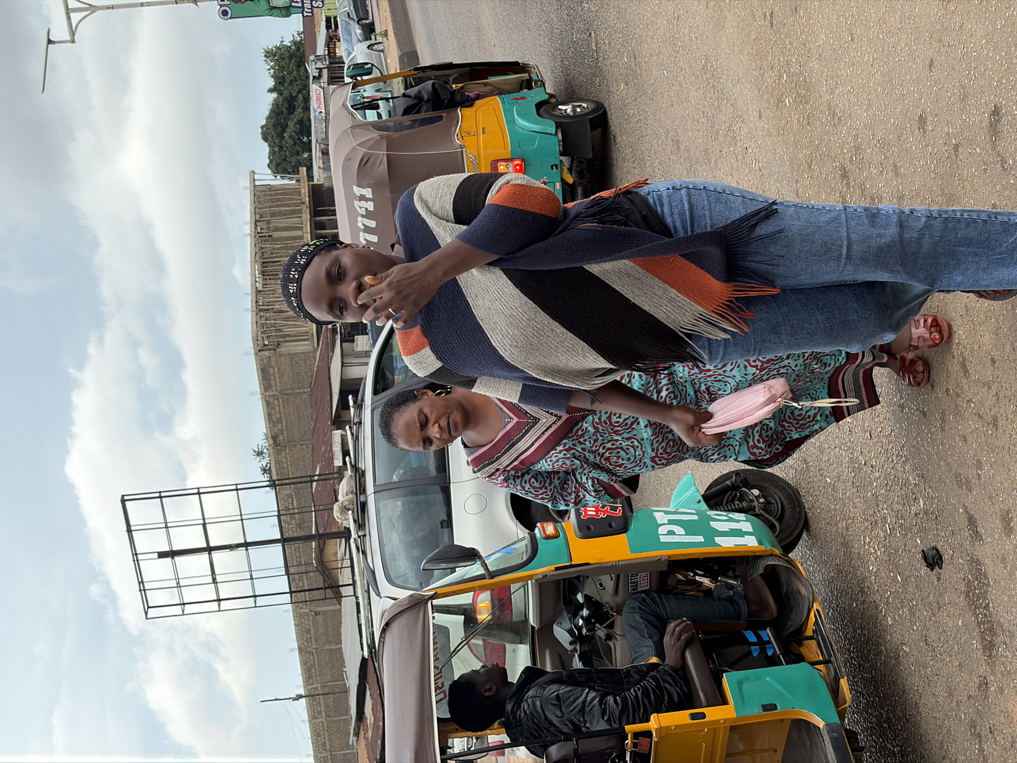 An outdoor, medium-angle, eye-level shot captures a street scene in Gura Top, Nigeria. In the foreground, a woman in blue jeans and a striped poncho walks with a pink purse. She is holding a cigarette to her mouth. Behind her, another woman in a patterned dress walks with her. To the left, a young man wearing a black jacket sits in a yellow and green auto-rickshaw, which has the letters "PTA" and the number "112" visible. In the background, other auto-rickshaws, cars, and buildings are visible under a cloudy sky. The ground is paved with asphalt.