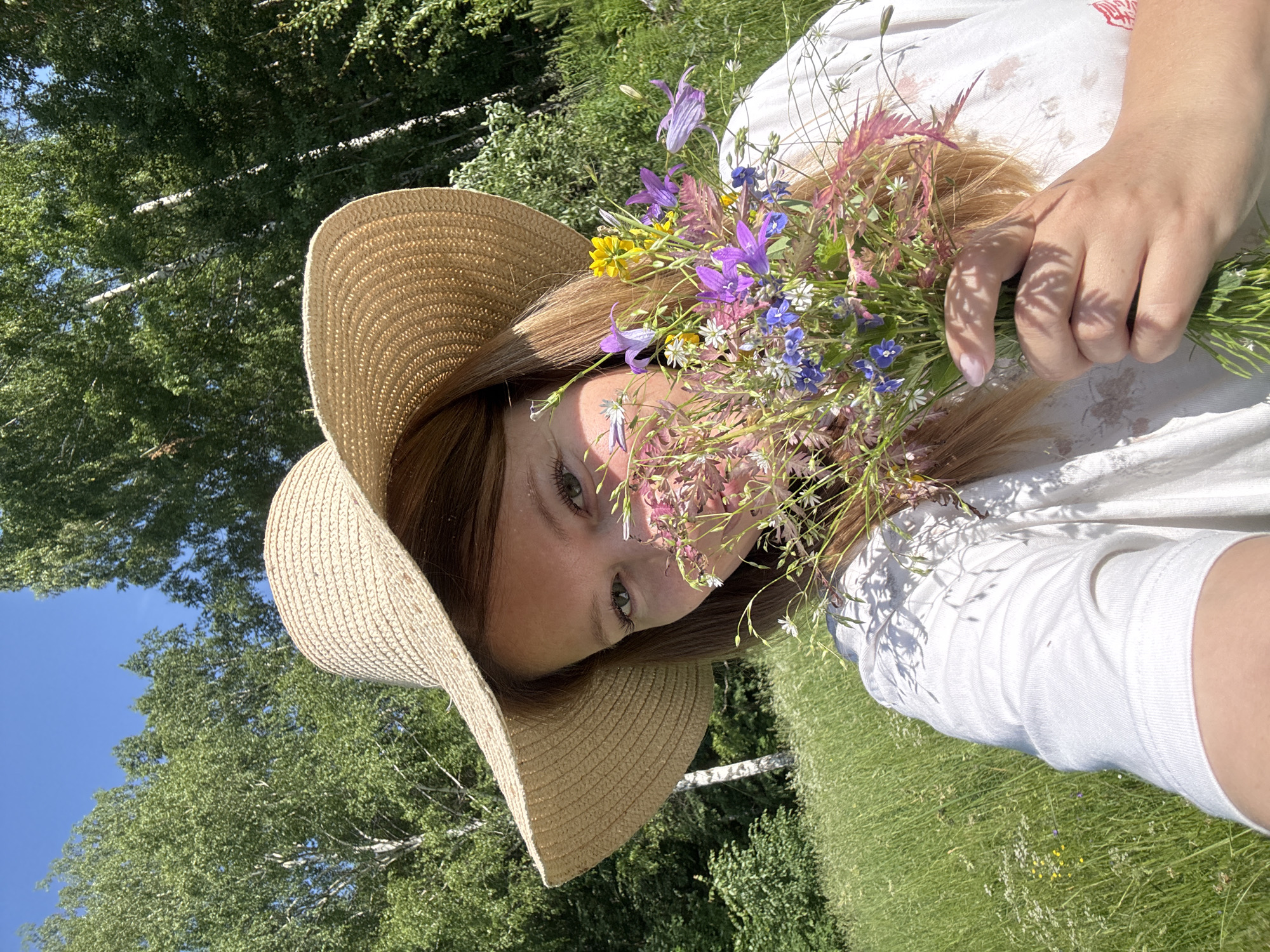 A young woman with blonde hair, wearing a wide-brimmed straw hat and a white shirt, is holding a bouquet of wildflowers in her right hand. She is partially obscured by the bouquet, with only her eyes and the side of her face visible. She is standing in a sunlit meadow with tall green grass and scattered wildflowers, with a backdrop of trees. The sky is clear and blue. The scene appears to be in the countryside, potentially Vassyata, Russia, given the typical landscape.