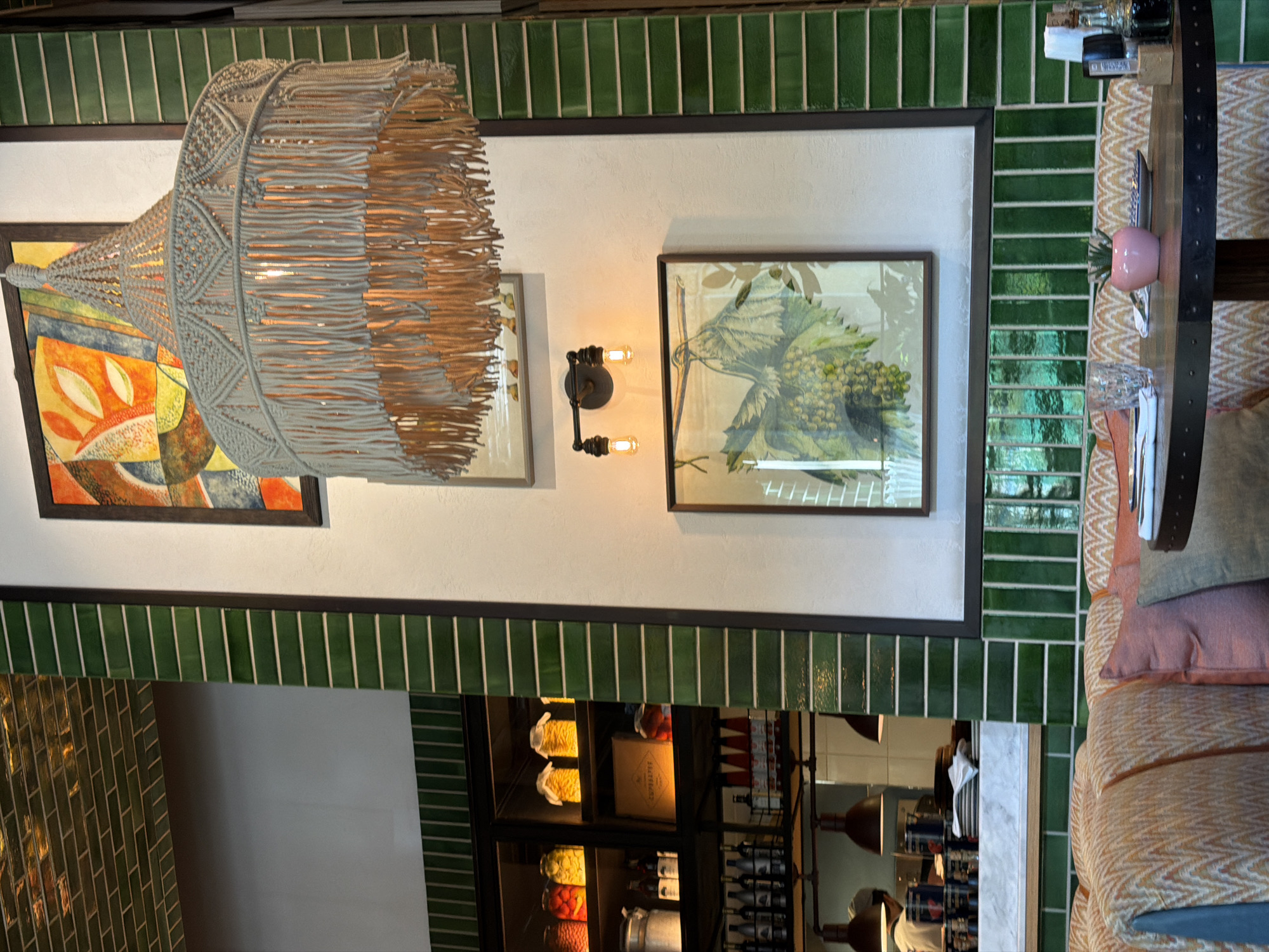 The image depicts the interior of a restaurant in Sabail Raion, Azerbaijan. The scene features a dining table in the foreground with upholstered seating. Above the table, a large, macrame-style pendant light with fringe hangs from the ceiling. The walls are adorned with framed artwork, including a still life of grapes and a more abstract piece. Green, rectangular tiles line portions of the walls and a shelf unit filled with jars and bottles is visible on the left. The overall ambiance suggests a contemporary and somewhat rustic dining establishment.