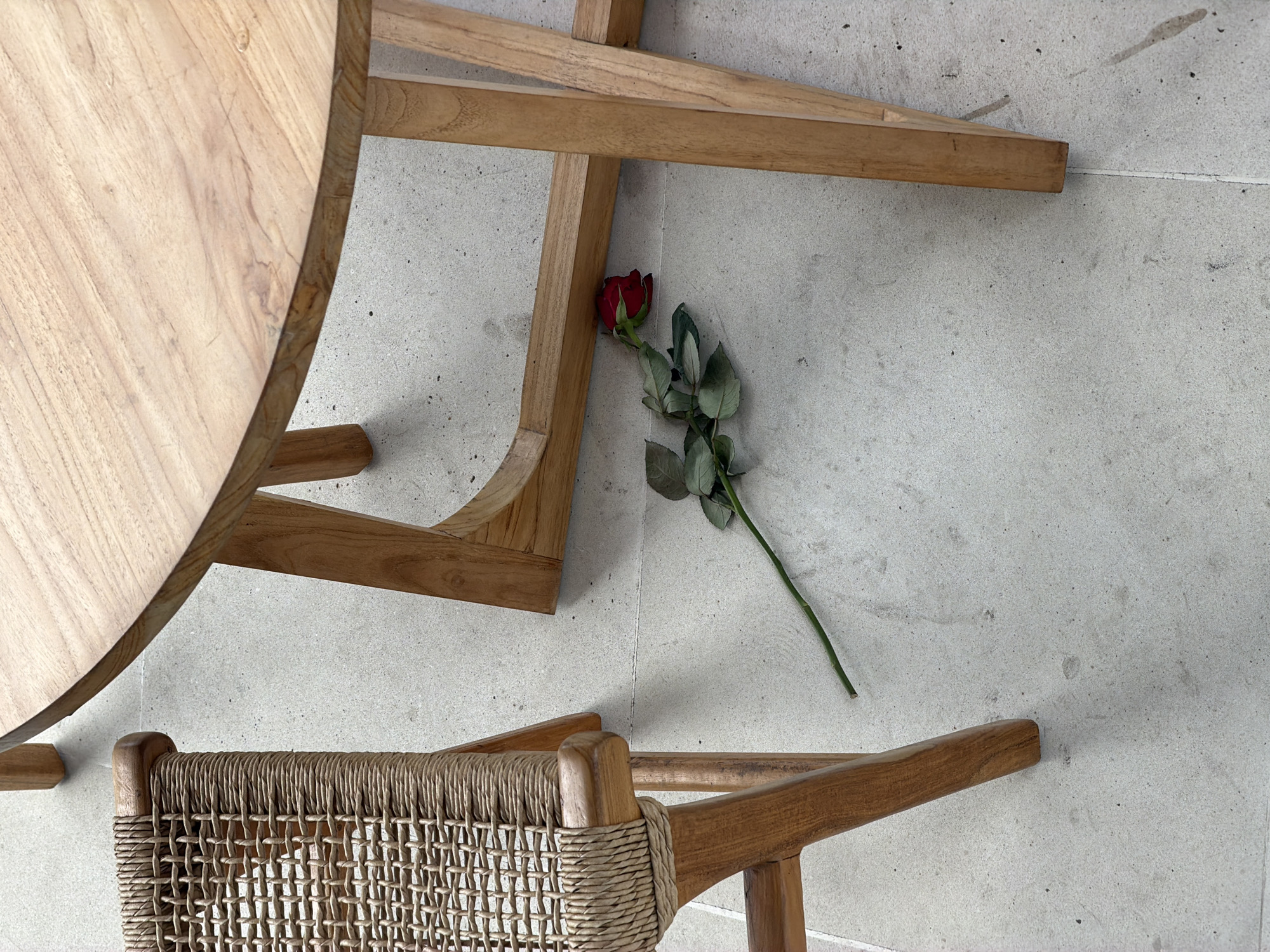 The image shows a top-down view of a sparsely decorated scene. A round, light-wood table occupies the left side of the frame. Next to the table, on the lower portion of the image, sits a chair with a woven back. A single red rose with green leaves and a stem lies diagonally between the table and the chair. The background is a pale, textured surface. The location is Mengwi, Indonesia.