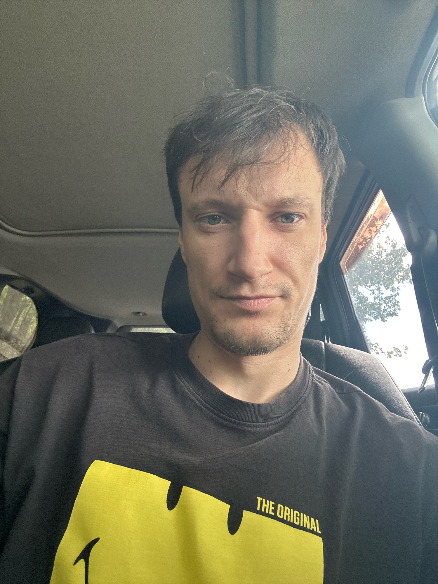 A man with short, messy brown hair and light blue eyes is shown from the chest up, inside a car. He is wearing a black t-shirt with a large yellow smiley face graphic and text that reads "THE ORIGINAL". The man is looking directly at the camera, with a neutral expression. The background shows the interior of the car, including the headliner, a sunroof, and a window with trees visible outside. The lighting suggests it is daytime. The image does not provide enough information to determine the specific location within Makiv, Ukraine.