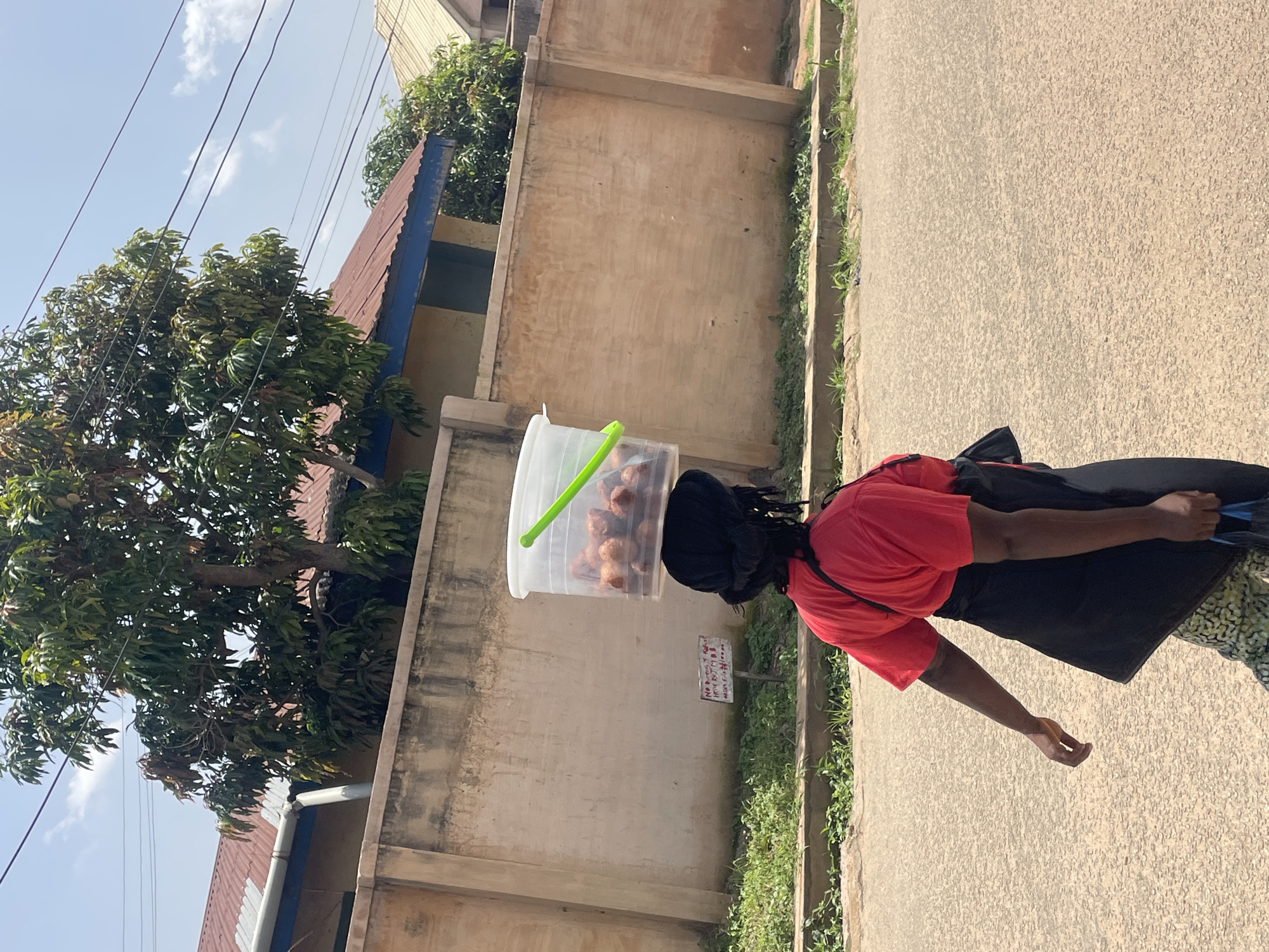 A woman walks on a paved road in the city of Jos, Nigeria, carrying a bucket of food on her head. The bucket is clear plastic with a green handle and contains several brown, spherical food items. The woman is wearing a red t-shirt, a black apron, and patterned pants. She has dark hair tied back and a dark purse slung over her shoulder. Behind her, a beige concrete wall runs along the side of the road, with a tree and a building visible above it. The sky is blue with scattered clouds. A small sign is posted on the wall in front of the woman.