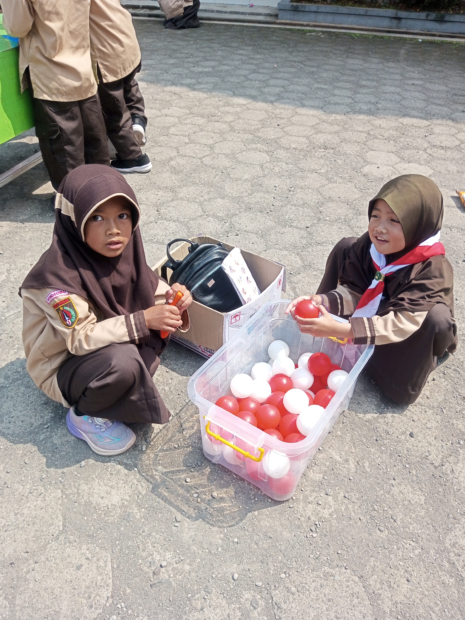 Two young girls in brown scout uniforms, one wearing a dark brown hijab and the other a lighter brown hijab, are sitting on a paved ground. The girl on the left, looking directly at the camera, is holding an orange object. The girl on the right, with a smile, is holding a red ball. In front of them is a clear plastic container filled with red and white balls, and a cardboard box with a black bag inside. The background shows a partially visible person in a similar uniform and a paved area with cracks. The scene appears to be outdoors, possibly a schoolyard or public space in Kalibeber, Indonesia, indicated by the context provided.
