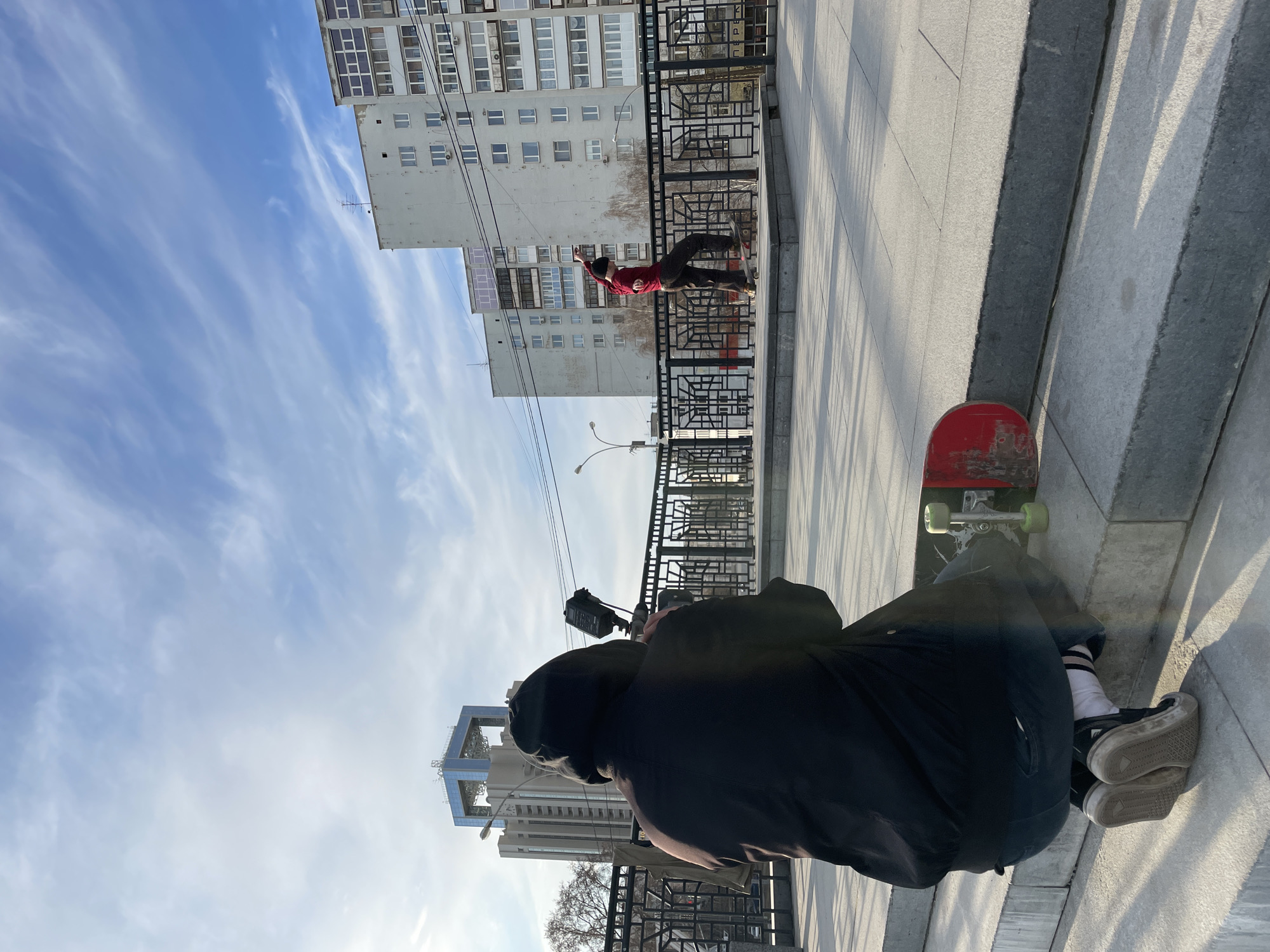 A person in black clothes and light-colored sneakers sits on the ground with their back to the viewer, holding a camera pointed towards a skateboarder performing a trick on a ledge. The skateboarder is wearing a red shirt and dark pants. A red skateboard is positioned next to the person with the camera. The background includes buildings and a partly cloudy sky. The location appears to be in Yekaterinburg, Russia, as evidenced by the architecture of the buildings in the background, including the "Iset" hotel.