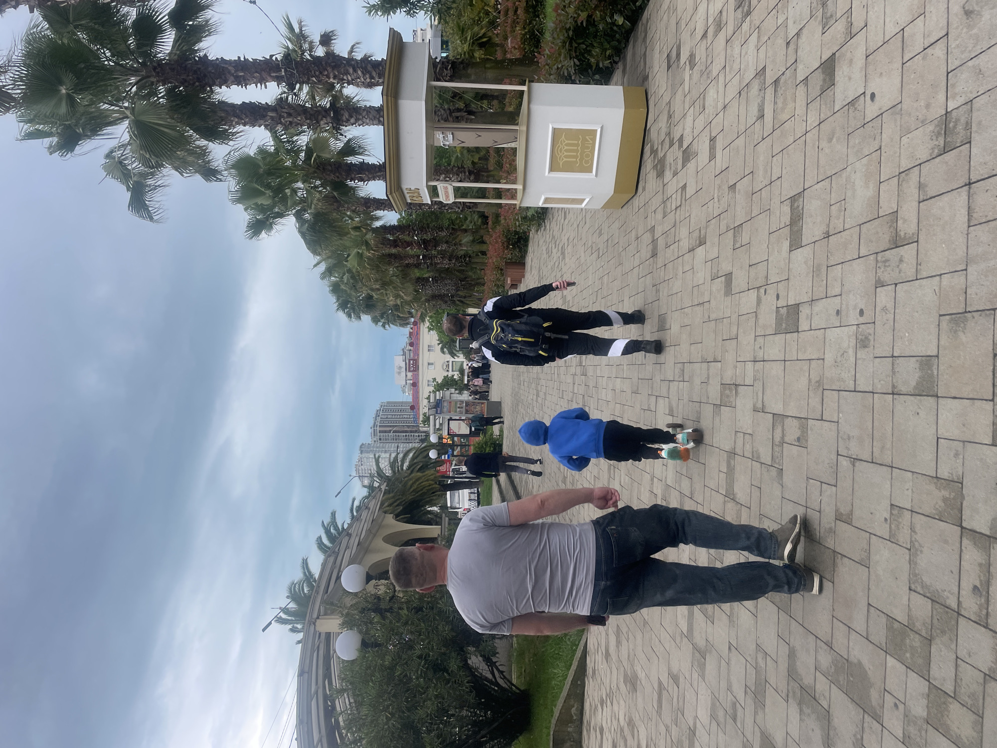 The image depicts a street scene in Sochi, Russia, as indicated by the "Сочи" sign on a ticket booth-like structure. The environment includes palm trees, other trees, modern buildings in the background, and paved walkways. The sky is overcast.
There are several people visible:
- In the foreground, a man is walking with his back to the camera, wearing a grey t-shirt and dark pants.
- Further along the walkway, a child is skateboarding.
- Another individual is walking away from the camera wearing a black and white jacket.
- Additional people are visible in the background, appearing as pedestrians.
Actions and interactions observed include walking, skateboarding, and general street activity. The scene is a typical urban pedestrian environment.