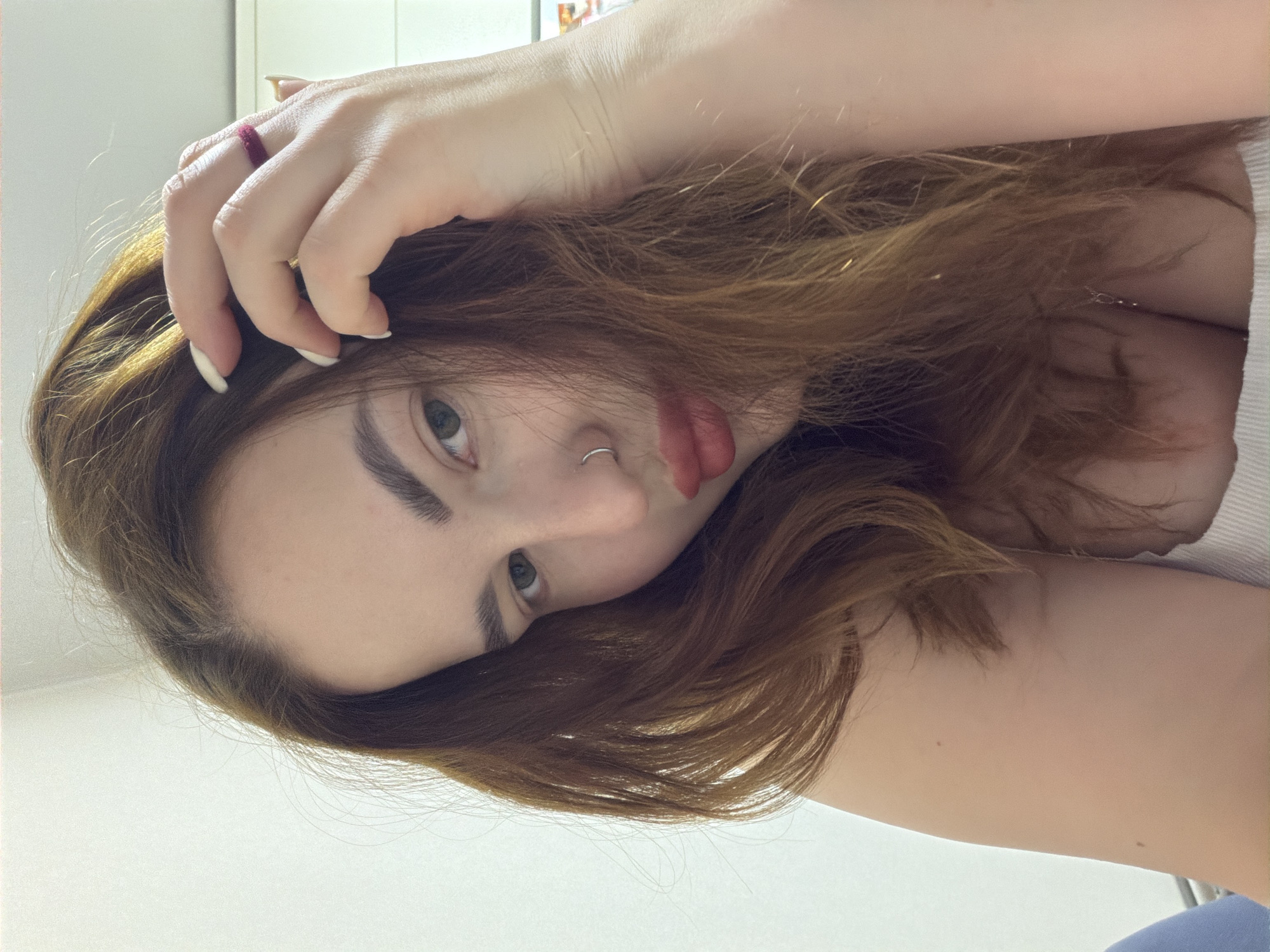 A close-up, vertically oriented portrait features a young woman with light brown to auburn hair, captured in an indoor setting in Krasnodar, Russia. Her head is tilted, and her direct gaze meets the camera. She has distinct green or hazel eyes, dark, prominent eyebrows, and full lips with a reddish-pink hue. A silver nose ring is visible on her right nostril.
Her right hand is positioned near her head, with fingers loosely entangled in her hair. A small, dark red ring adorns her left ring finger, and her nails are manicured with white tips. She appears to be wearing a light-colored, possibly white, ribbed top, partially visible on the right side of the frame. A glimpse of blue fabric or an object is present in the lower right corner. The background is simple, light-colored, and out of focus, suggesting a room with plain walls. The lighting is bright and even, indicative of daytime. Her expression is serious and contemplative. There is no visible text.