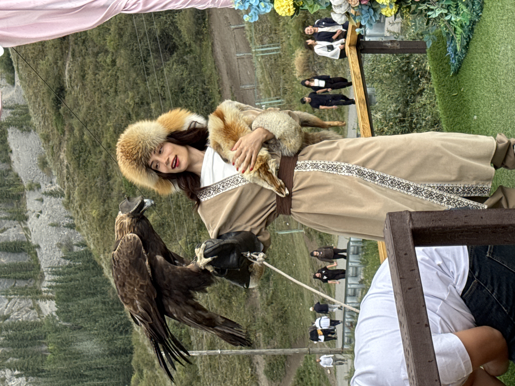 A woman wearing a traditional Kazakh outfit and a fur hat stands outdoors, holding a golden eagle on her gloved forearm. She is wearing a tan robe with intricate white trim, a brown belt, and a white shirt. She holds a piece of fur in her left hand. The eagle wears a leather hood. In the background, a lush, green hillside is covered in trees and rocky outcrops. Several people are visible walking on a path behind a wooden railing. The scene appears to be taking place in the city of Kormeti, Kazakhstan, likely a tourist attraction.