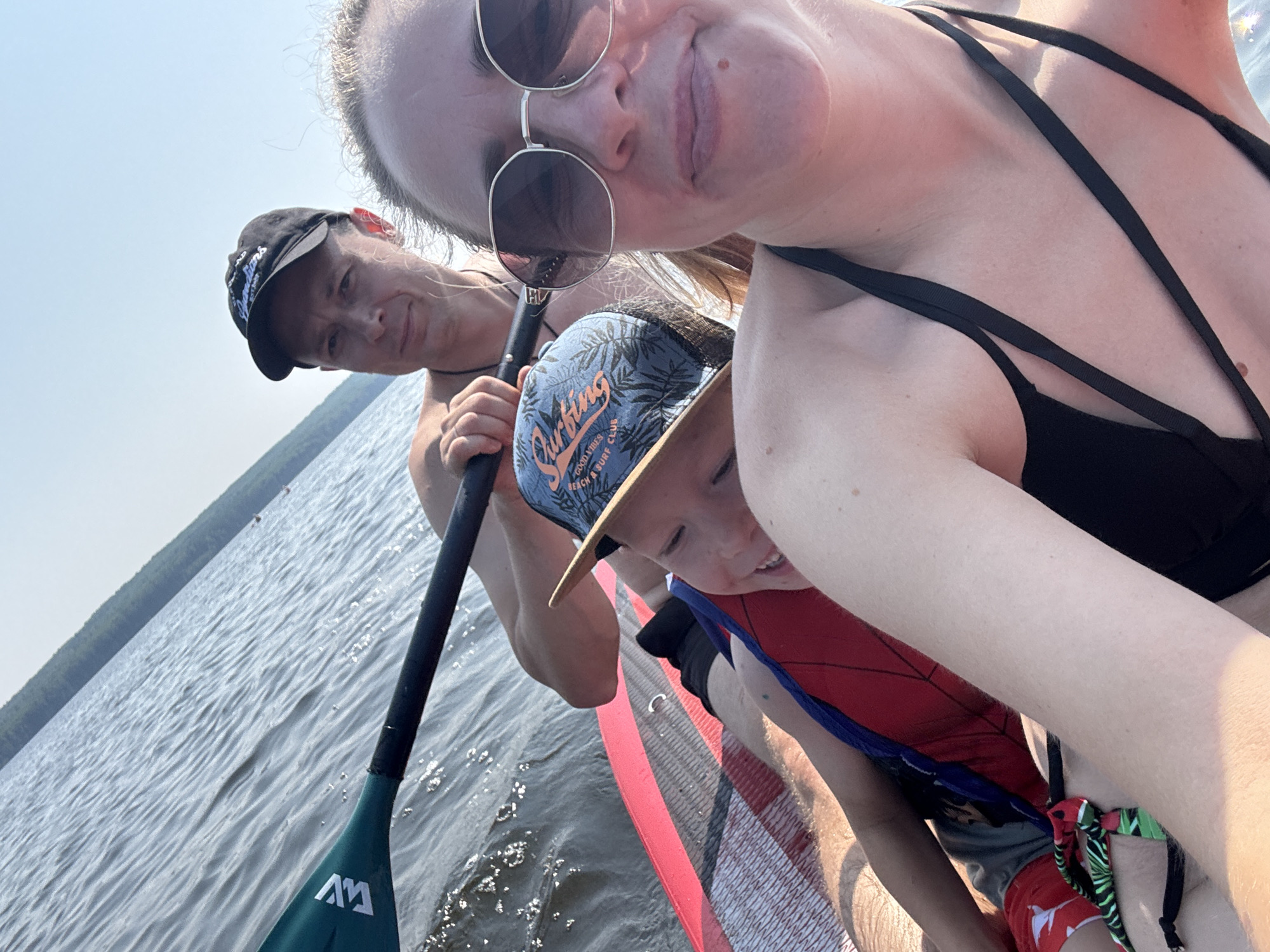 An outdoor, eye-level, full selfie shows a family of three on a paddleboard in the city of Chaikovsky, Russia. In the foreground, a smiling woman with blonde hair, wearing sunglasses and a black bikini, looks at the camera. To her left, a young boy with blonde hair, wearing a blue baseball cap and a red life vest, is lying on the paddleboard. Behind him, a man with brown hair and a black baseball cap is holding a paddle. The family is on a body of water with a tree-lined shore in the distance. The sky is clear and blue.