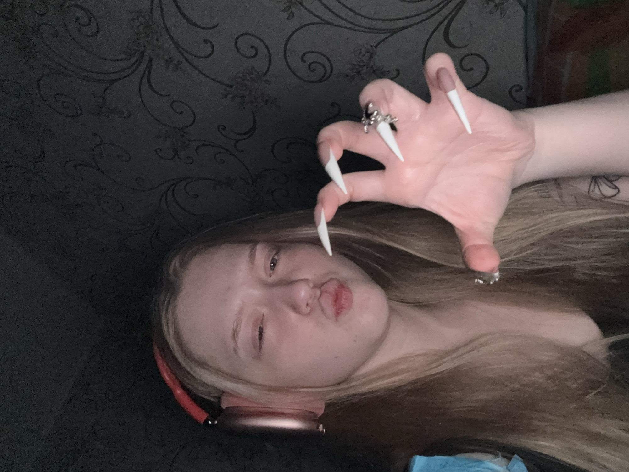 A young person with blonde hair, wearing pink headphones, is lying down. They are making a kissing face towards the camera and holding up a hand with long, white, stiletto-shaped false nails and silver rings. The background is a dark gray, patterned wall. The location is unspecified, and there is no information to indicate the city of Saratov, Russia.