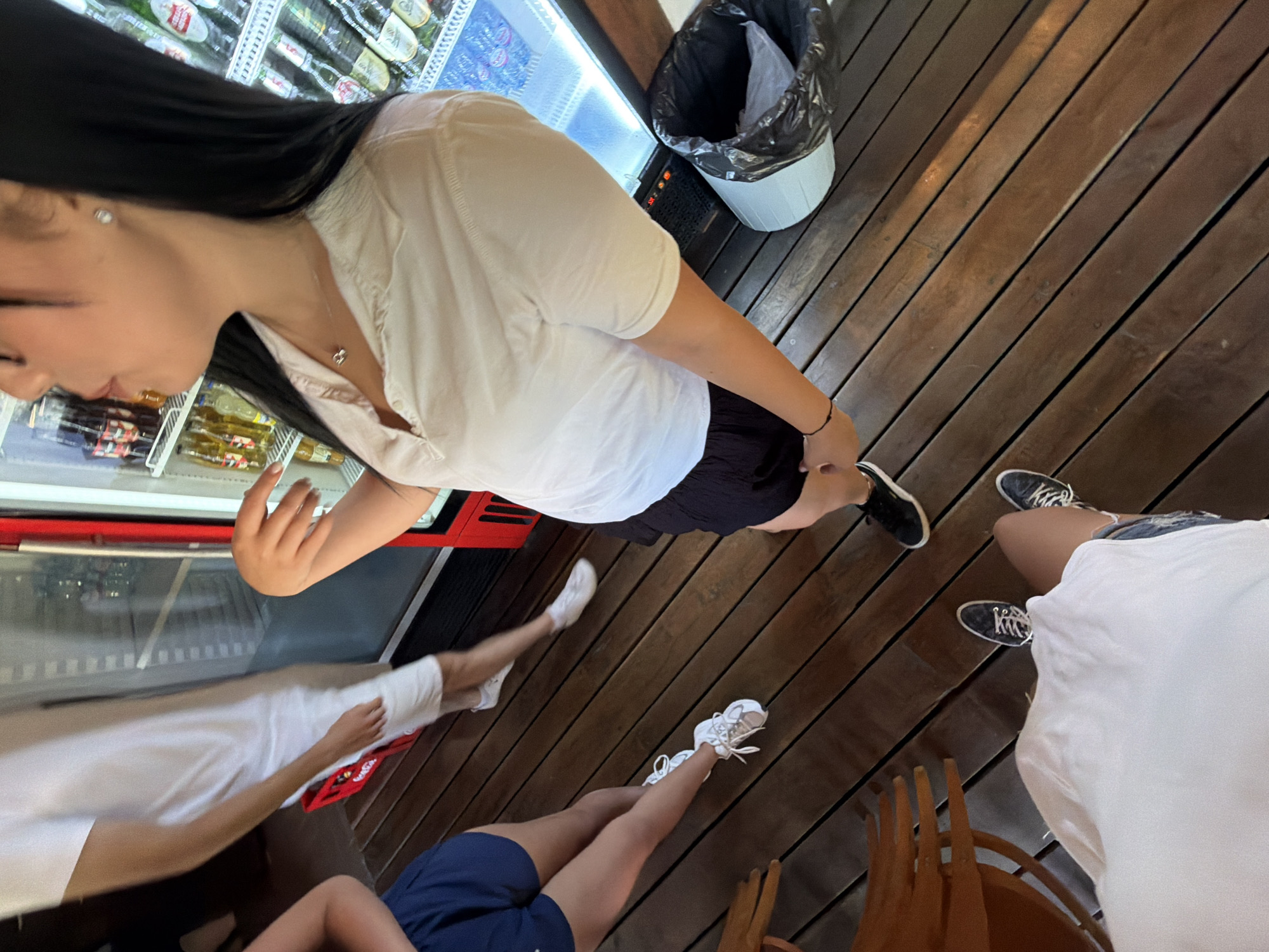 This image captures a casual outdoor scene, likely at a bar or cafe, on a wooden deck in Tsarevo, Bulgaria. In the foreground, a person wearing a white t-shirt and denim shorts is seated, their legs visible. To their left, two other individuals are standing. One is wearing navy blue shorts and a white t-shirt, and the other is wearing white shorts and a white t-shirt. In the background, a large red refrigerator stocked with beverages is visible. A black trash can with a light blue liner is situated next to the refrigerator. The overall atmosphere suggests a relaxed, possibly summer gathering.