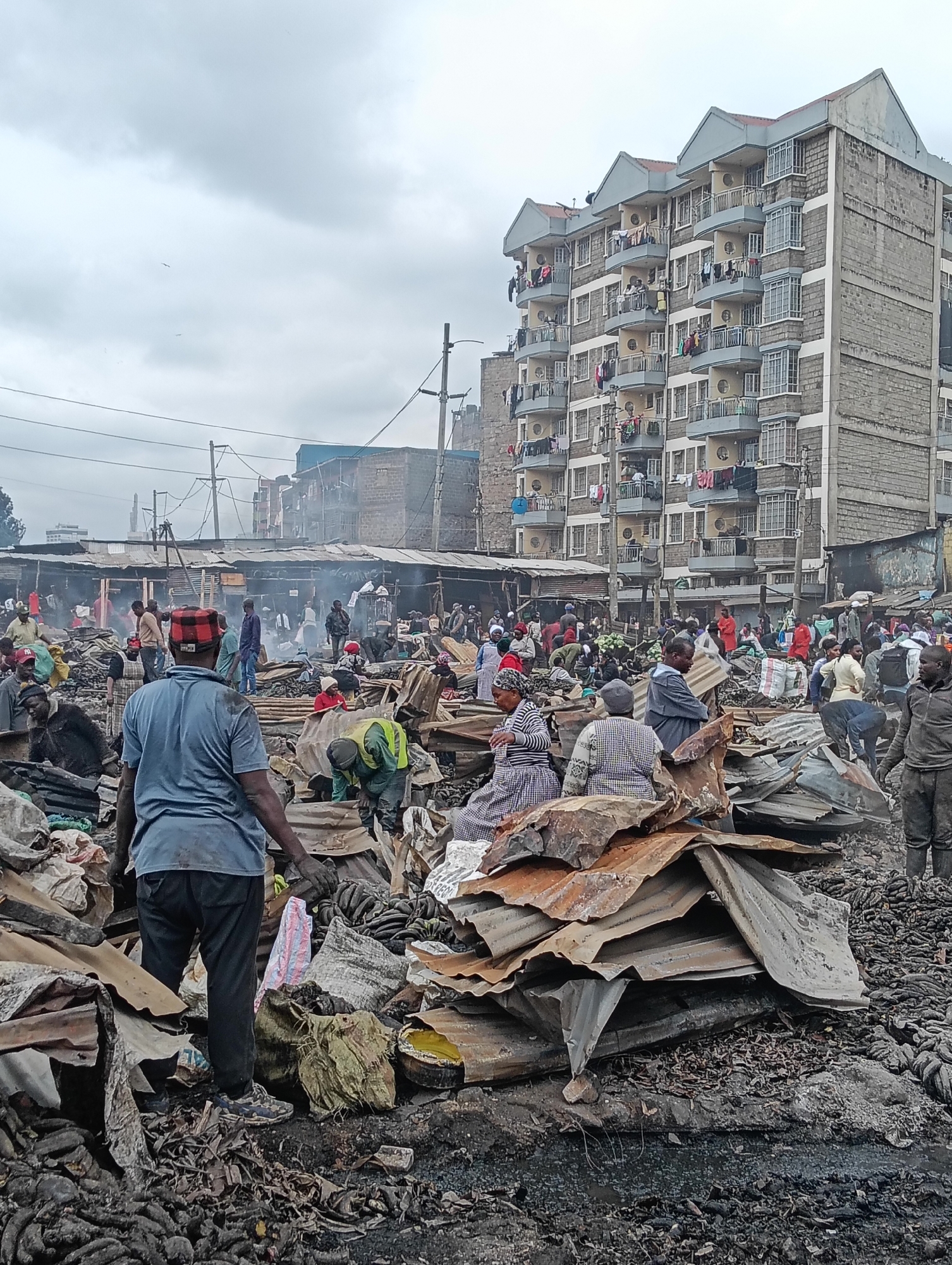 This is a ground-level, medium-shot photograph of a market in Nairobi, Kenya, following what appears to be a destructive event such as a fire or demolition. The scene is chaotic, with piles of debris, including corrugated metal sheets, charred materials, and scattered produce like bananas, dominating the foreground and mid-ground. Several individuals are present, some appearing to salvage items from the wreckage, while others observe the scene.
In the background, a multi-story apartment building stands, contrasting sharply with the devastation in the market. The building has balconies with laundry hanging out to dry, suggesting ongoing life in the residential area despite the market's condition. The sky is overcast, contributing to a somber atmosphere.
The people visible are of varying ages and genders, dressed in casual clothing. Their activities range from searching through the debris to standing and conversing. The overall impression is one of displacement and recovery in a dense urban environment. The presence of electricity poles and overhead wires indicates the market is situated within a developed area of the city.
