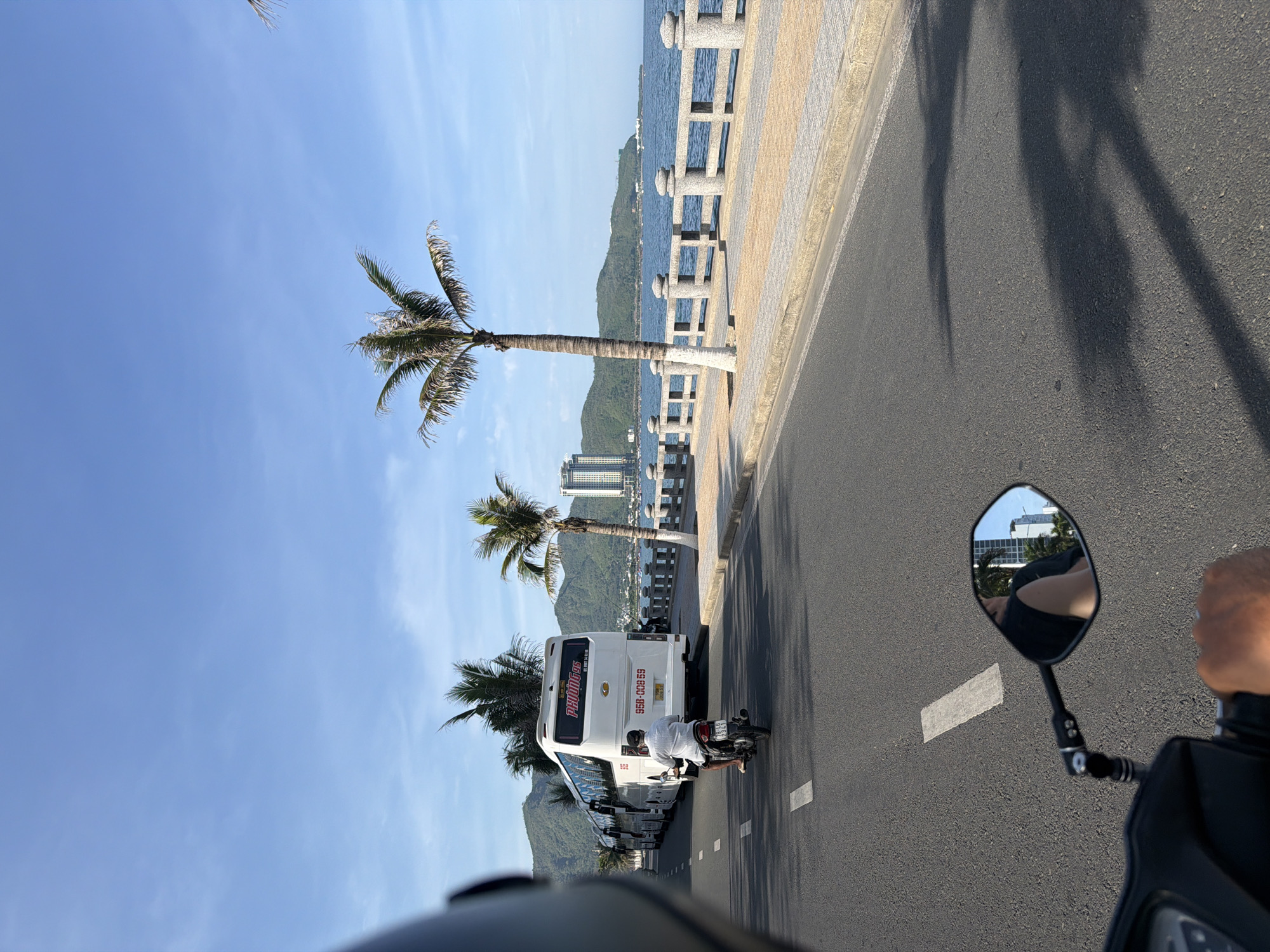 A frontal view from a motorcycle shows a road alongside a coastline. A line of white tour buses is parked on the right side of the road, with a single motorcycle rider in front of them. Beyond the buses, there is a stone walkway with a railing, followed by a body of water with a distant shoreline featuring mountains and a tall building. Palm trees are present along the walkway. The sky is clear and blue.