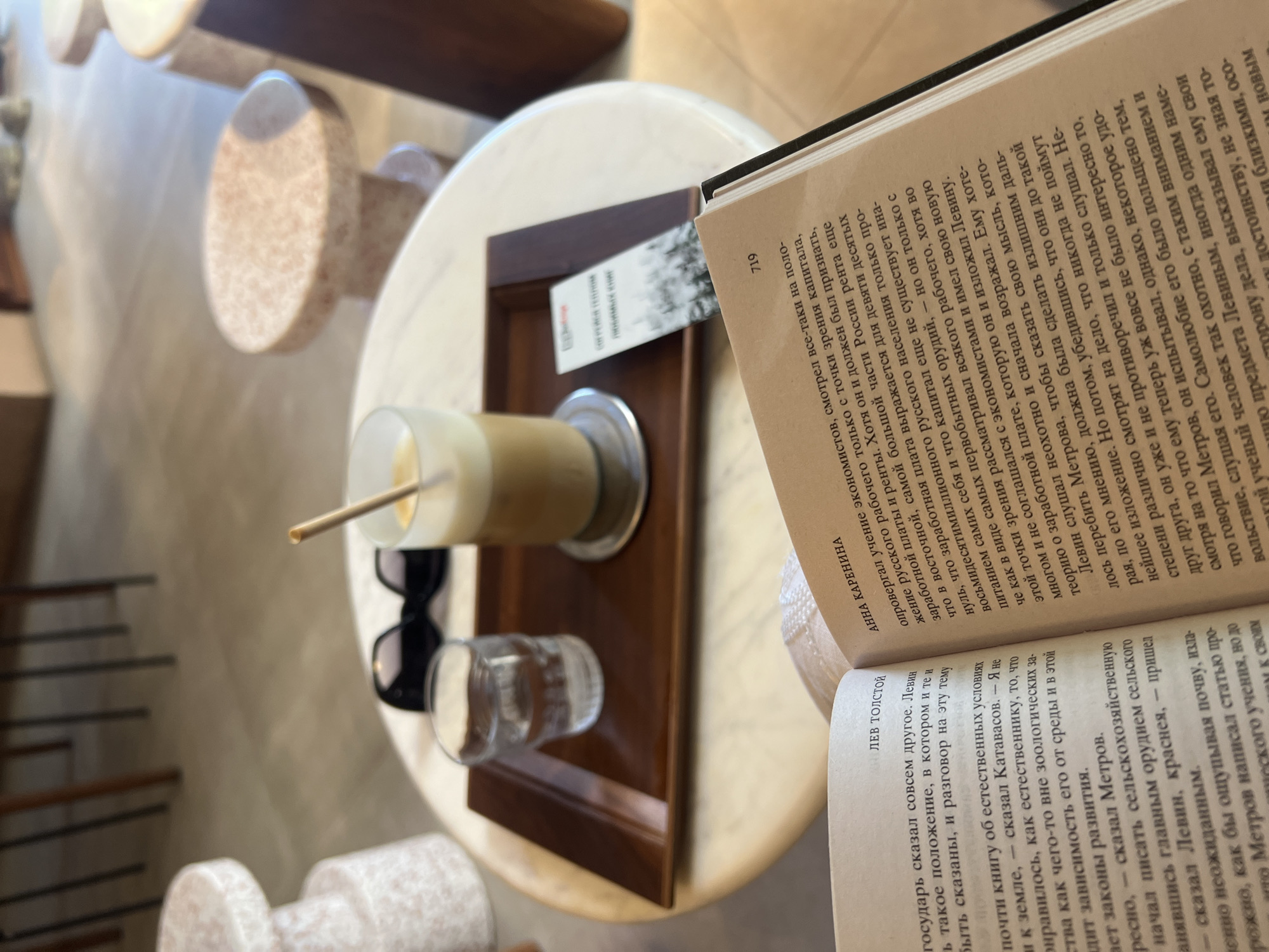 The image shows a close-up, eye-level view of a marble-topped table in what appears to be a cafe or outdoor seating area. On the table rests an open book, a glass of water, a glass with a frothy beverage and a straw, and a pair of black sunglasses. A small, dark wooden tray holds the two glasses. The book's pages are open, with text visible, and a bookmark with an image and text is tucked into it. The book is identified as "Анна Каренина" by ЛЕВ ТОЛСТОЙ, with page number 719 visible. The beverage appears to be a coffee drink, possibly an iced latte or cappuccino. In the background, out of focus, are parts of what seem to be light-colored, textured stools and furniture, suggesting a modern or minimalist aesthetic. The lighting is soft, indicating it might be daytime, possibly with natural light. The scene is static and lacks any visible people or active interactions, conveying a sense of quiet contemplation or relaxation. There is no indication that this scene is located in Barcelona, Spain.