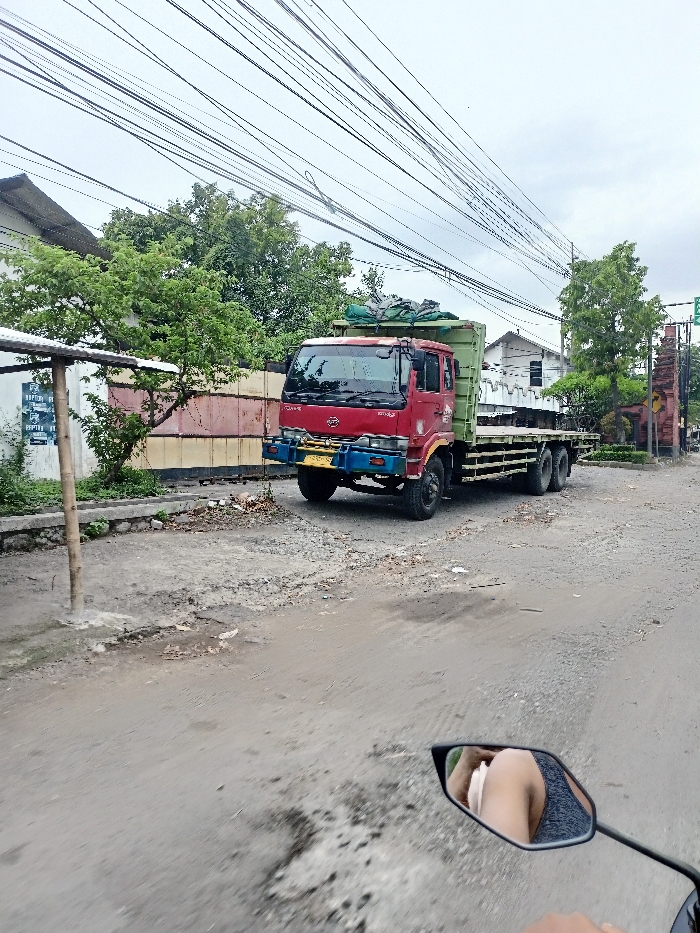The image captures a street scene in Kecamatan Mojosari, Indonesia, from the perspective of someone on a motorcycle, indicated by the reflection of an arm and leg in a rearview mirror in the bottom right corner.
The primary subject is a large, red-cabbed flatbed truck with a green bed, possibly a dump truck, parked or stationary on the side of a dirt and gravel road. The truck features a multi-colored bumper, primarily yellow and blue. Overhead, numerous black power lines crisscross against a cloudy, light gray sky.
To the left of the truck, there are green trees and bushes, and a section of a fence or wall that appears to be made of corrugated metal, painted in shades of red and tan. A wooden post stands in the foreground on the left, next to a sign with partially visible white text that reads "RAPTOR RU". Further back and to the left, a white building with a dark roof is partially visible.
To the right of the truck, the road continues, and another section of green trees and bushes can be seen, along with what appears to be a decorative archway or gate structure made of brick or stone. The road surface is unpaved or in disrepair, with visible potholes and loose dirt/gravel.
The lighting suggests it is daytime, possibly under an overcast sky, as there are no harsh shadows. The overall scene depicts a typical, somewhat rustic street environment with utilitarian infrastructure.