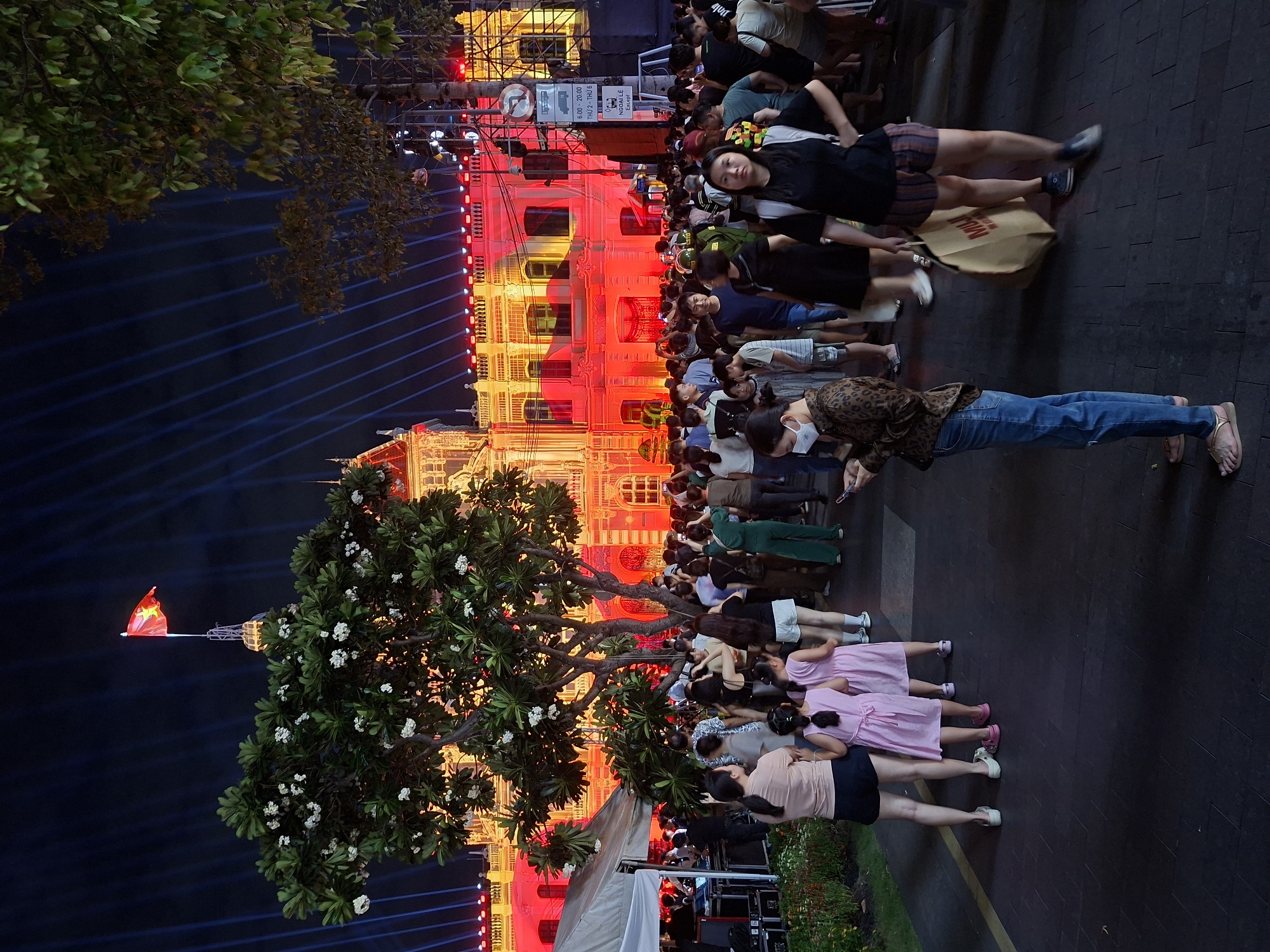 The image captures a nighttime scene in Thủ Đức, Vietnam, likely during a public event or celebration. A large, ornate building, illuminated with vibrant red and yellow lights, serves as the backdrop. A crowd of people, diverse in age and attire, are gathered in front of it, suggesting a shared experience. Some individuals are looking towards the building, while others are interacting with each other.
In the foreground, a lush green tree with white blossoms adds a natural element to the urban setting. A Vietnamese flag is visible, flying high on a pole, hinting at a patriotic or official occasion. The sky is dark, with prominent blue light beams projecting upwards, creating a dramatic and festive atmosphere.
Among the crowd, several individuals are clearly visible. A woman in a leopard-print jacket and blue jeans stands out, as does a group of young girls in pink dresses. The text visible on a sign reads "6:00-20:00 THỨ 2 - THỨ 6" and "NGOẠI LỆ Except", likely indicating operating hours or special conditions, possibly related to the building or event. The overall impression is one of a lively and communal gathering under the glow of artificial lights and a clear night sky.