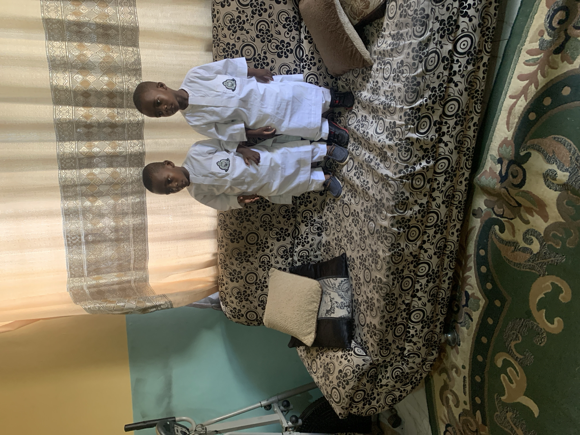 Two young boys, dressed in matching white traditional garments, are seated on a brown floral patterned couch in a room. The couch has two small pillows. Behind the couch, a beige and silver patterned curtain hangs in front of a wall painted partially yellow and partially teal. A piece of exercise equipment is visible near the base of the couch. Part of a green and brown patterned rug is visible on the right side of the image. Location is Gura Top, Nigeria.