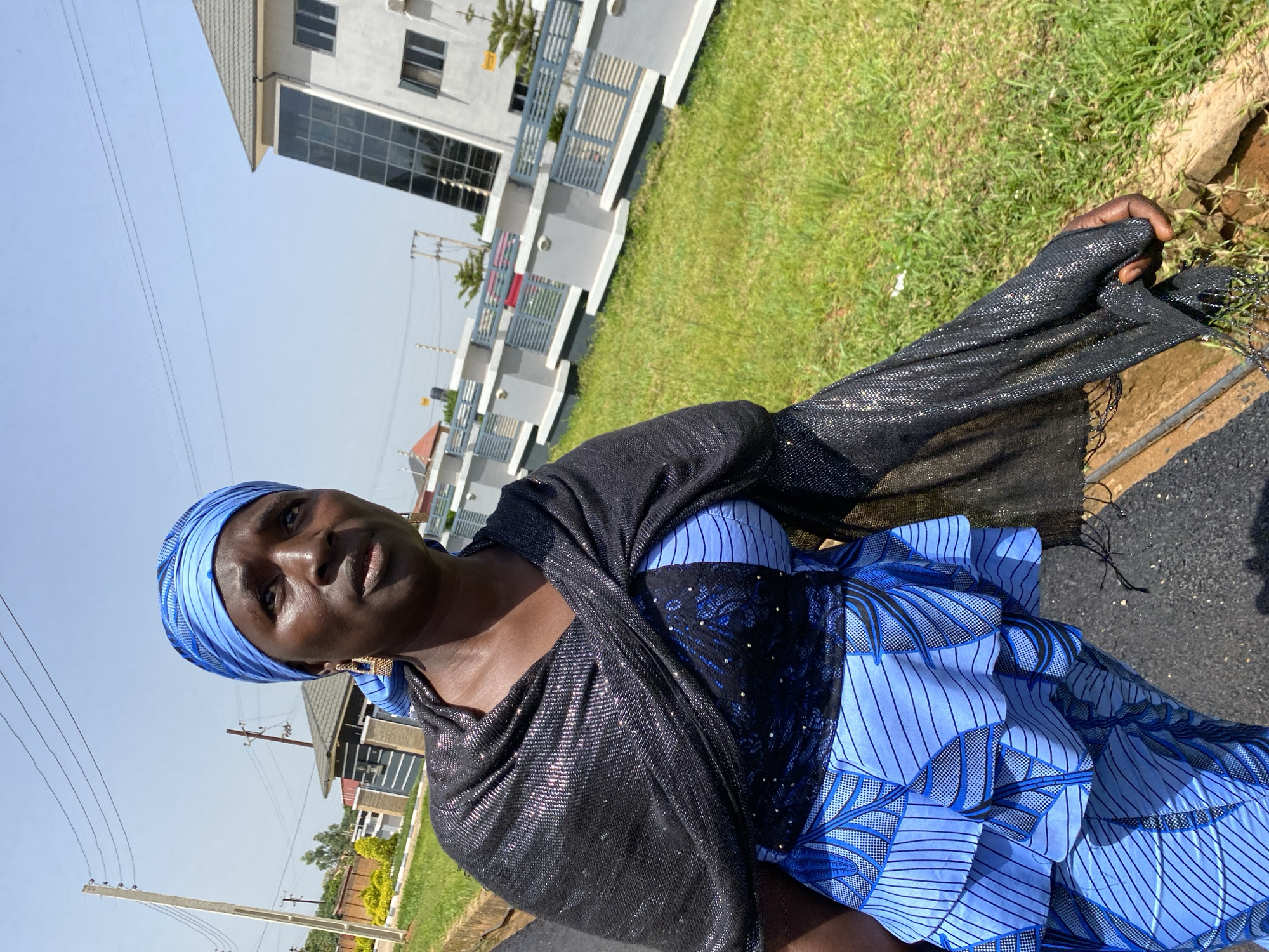 A woman is captured in a close-up, outdoor portrait, oriented vertically. She is wearing a blue headscarf and a blue and black patterned dress, draped with a sparkly black shawl. The woman looks towards the camera with a neutral expression. In the background, a modern white building with large glass windows is visible, along with a well-maintained grassy area and what appear to be other residential structures. Power lines and poles are also present in the mid-ground, suggesting an urban or semi-urban setting. The scene is illuminated by bright sunlight, indicating daytime, and the sky is clear and blue. The image appears to be taken in Bukuru, Nigeria.