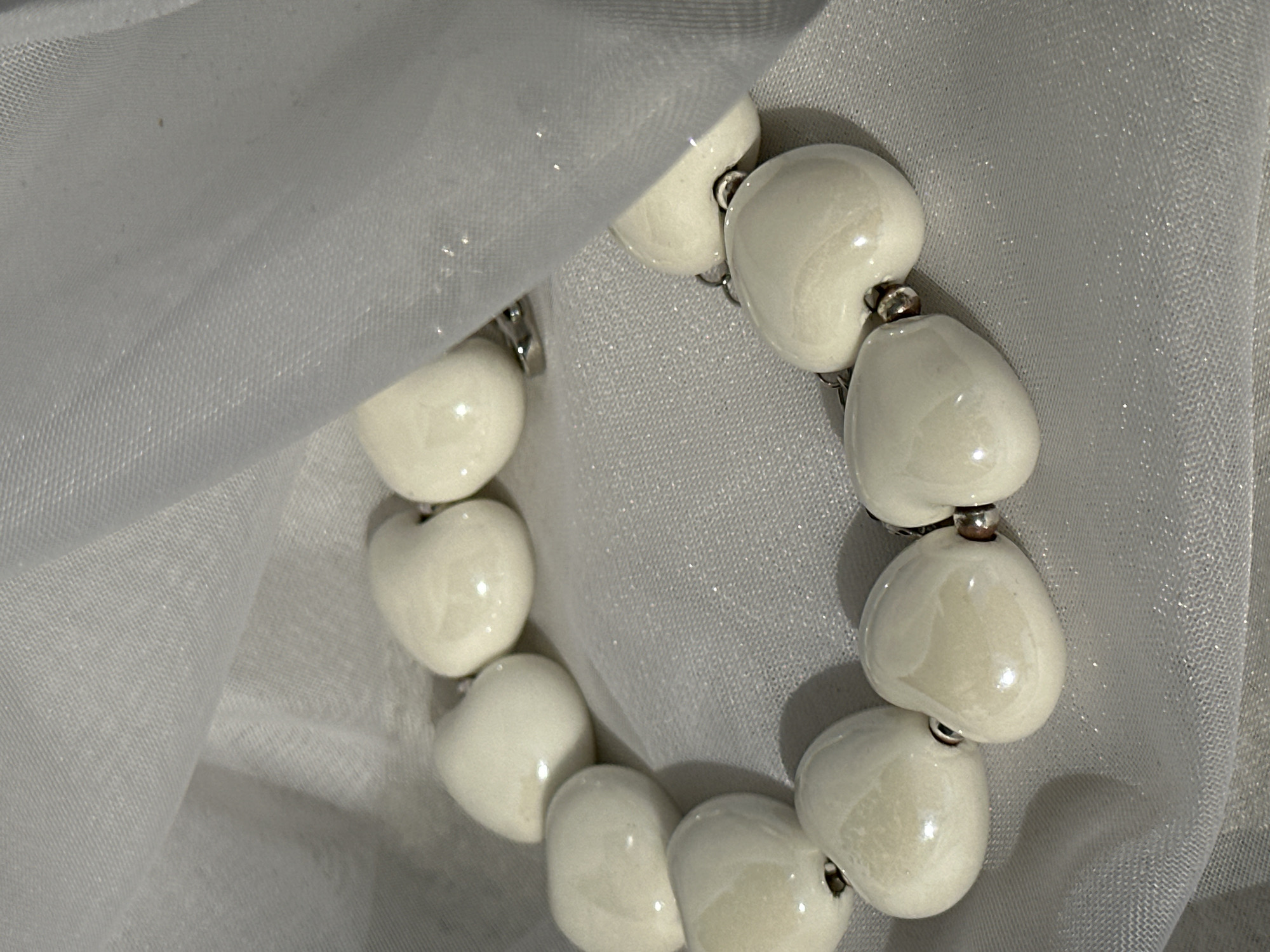 The image shows a close-up of a bracelet made of white, heart-shaped beads connected by small, silver-colored beads. The bracelet is resting on or draped with a piece of light-colored, semi-transparent fabric that has a slight sheen. No location is identifiable in the image.