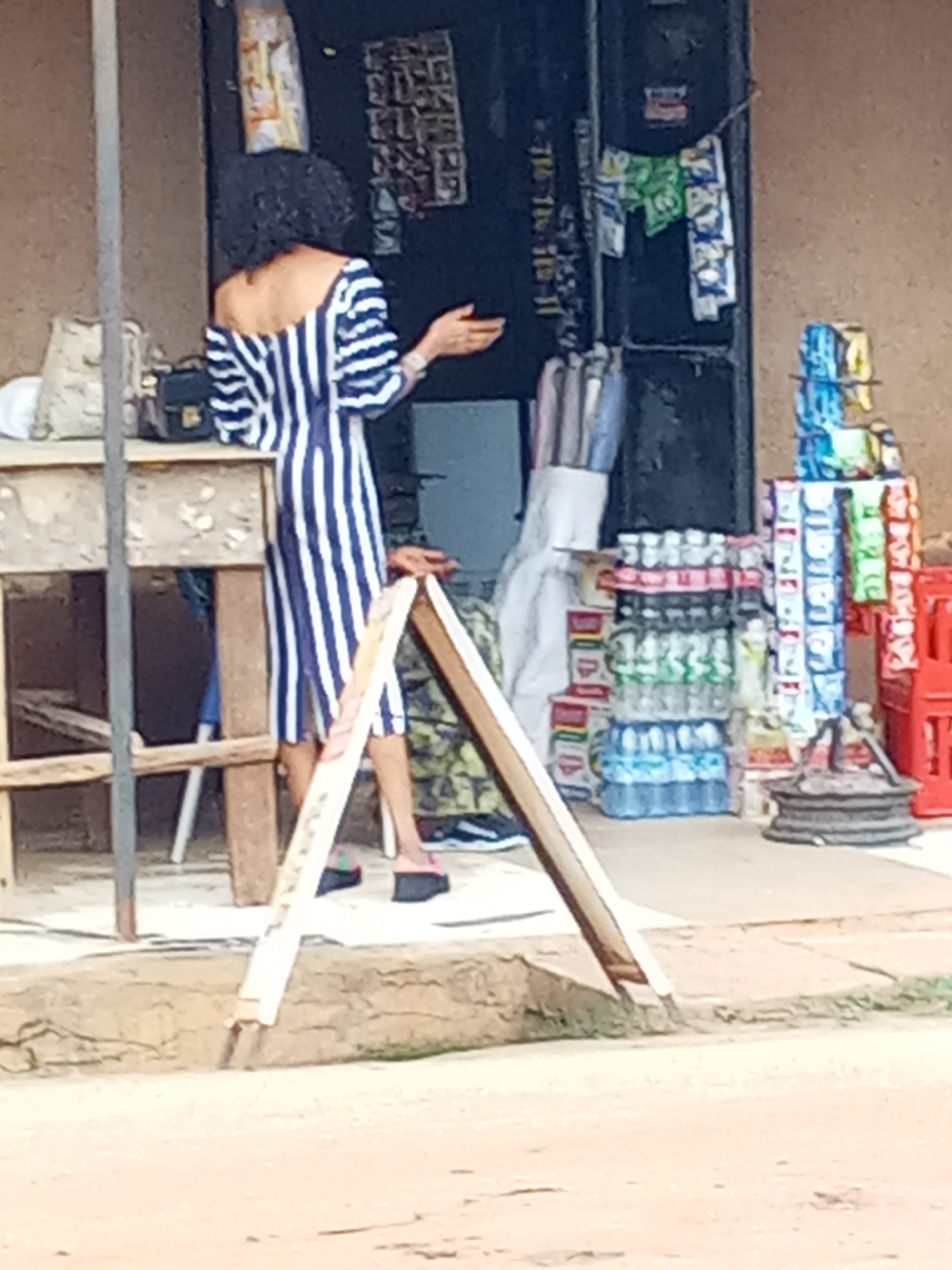 The image shows an open-air store in Sabon Tasha, Nigeria. A person wearing a blue and white striped dress is standing at the entrance of the store, gesturing with their right hand. The store sells bottled water, drinks in tetra packs, snacks, and other small items. A wooden table and chair are to the left of the person. An A-frame sign is partially visible in the foreground. The ground is a mix of dirt and pavement.