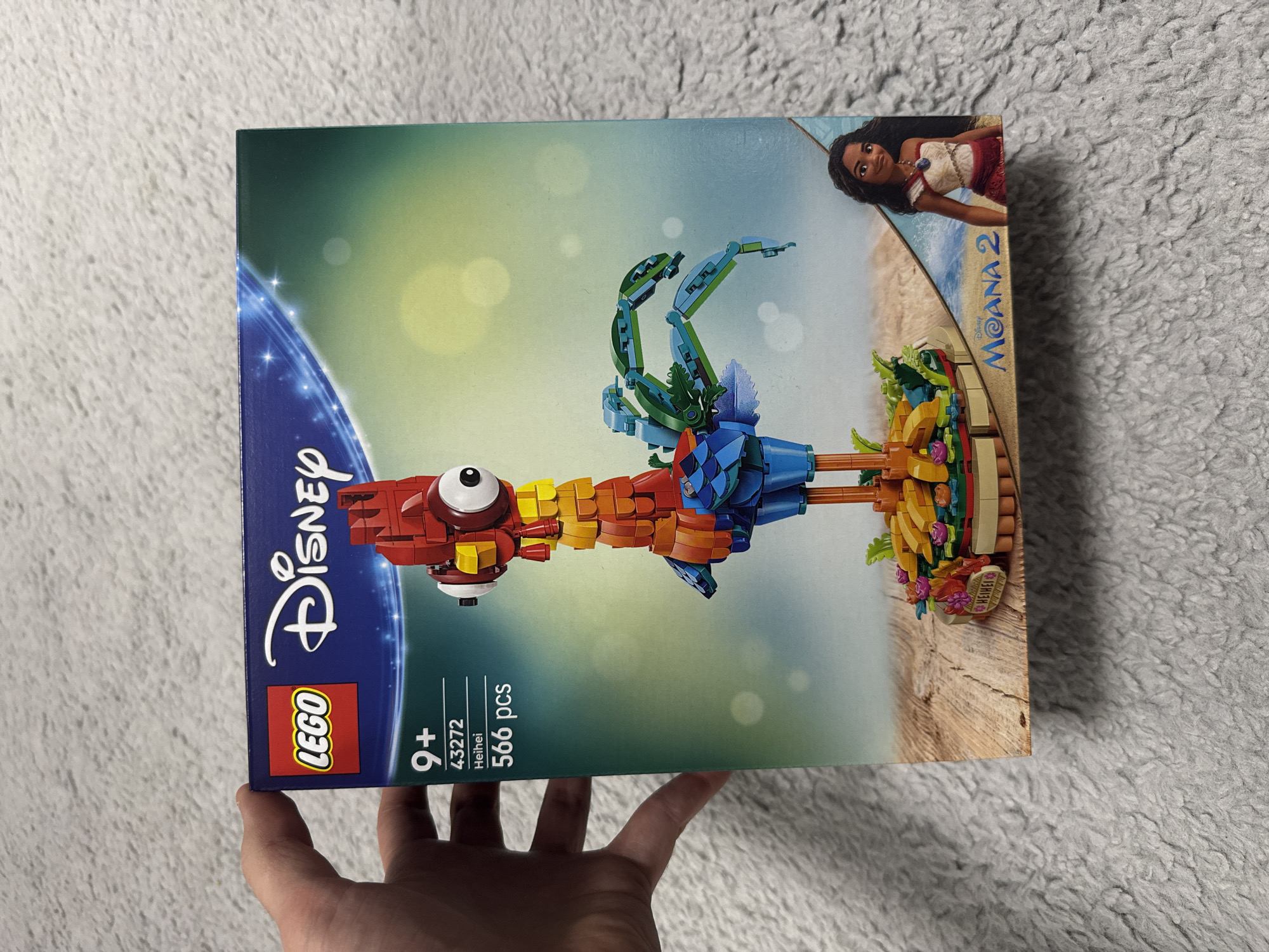 A close-up shot shows a person's hand holding a Lego Disney Moana 2 building set box, featuring a Lego representation of the character Heihei. The box is primarily green and blue, with depictions of Moana and the Lego Heihei against a beach backdrop. The LEGO logo and age recommendation (9+) are visible on the side of the box. The box is situated on a grey, textured surface. The location is not possible to determine from the image.